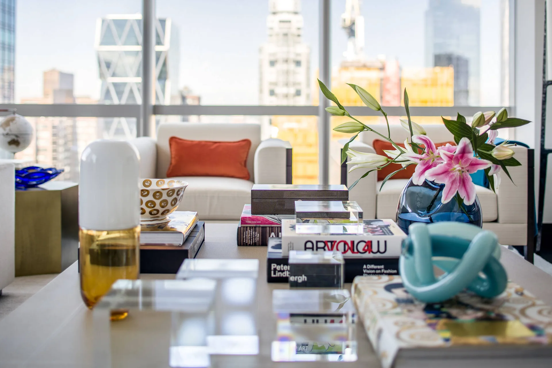 A close-up of a coffee table styled with stacked art books, a glass vase with lilies, and sculptures, with city buildings visible through windows.
