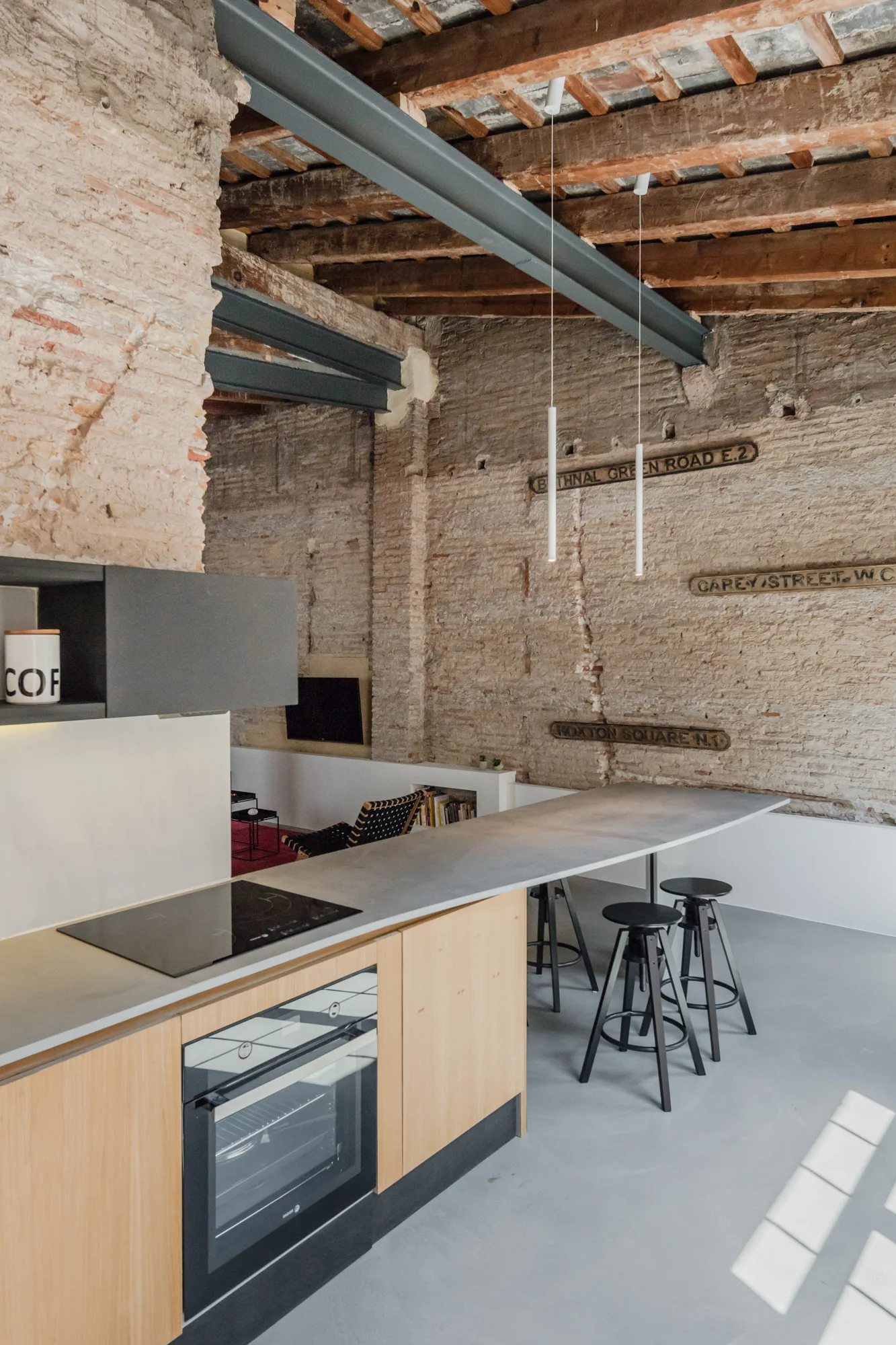 A modern industrial kitchen features a long concrete-look island with light wood cabinets, black stools, exposed brick walls, and timber ceiling beams.
