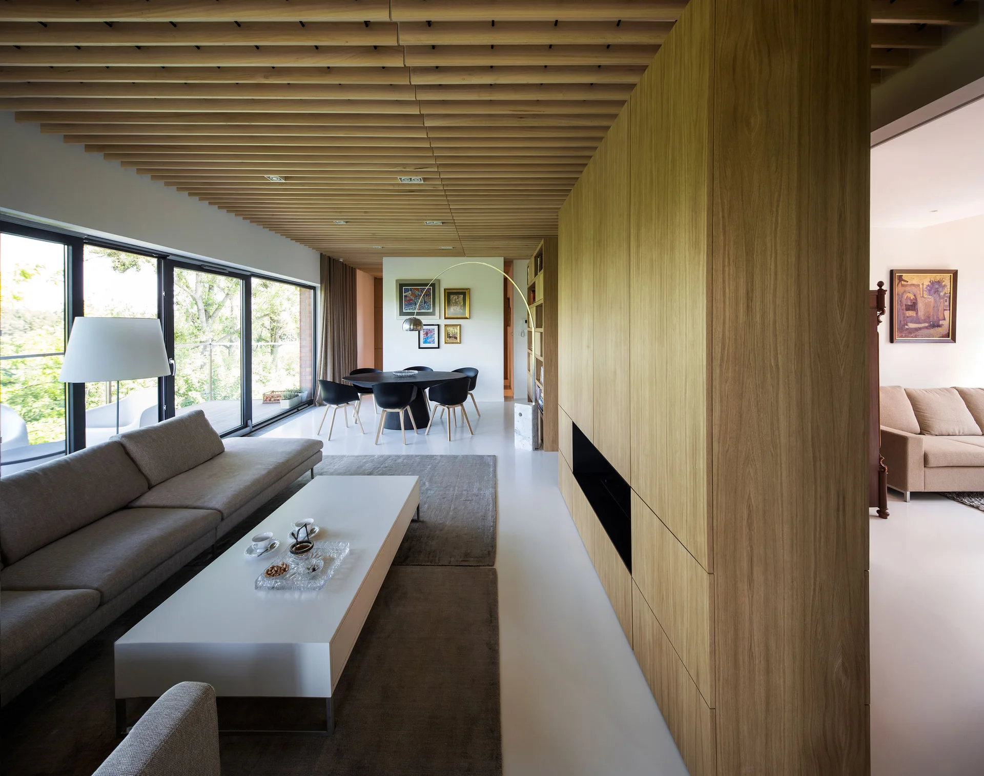 A minimalist living room features a wood-slatted ceiling, a large wood-grain storage divider wall, a grey sofa, and large sliding glass doors.