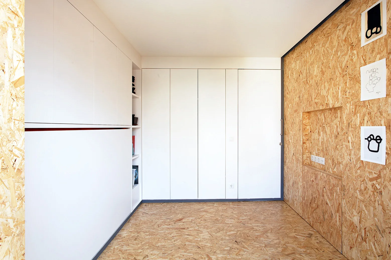Minimalist white floor-to-ceiling cabinets stand opposite a wall and floor covered in textured oriented strand board.