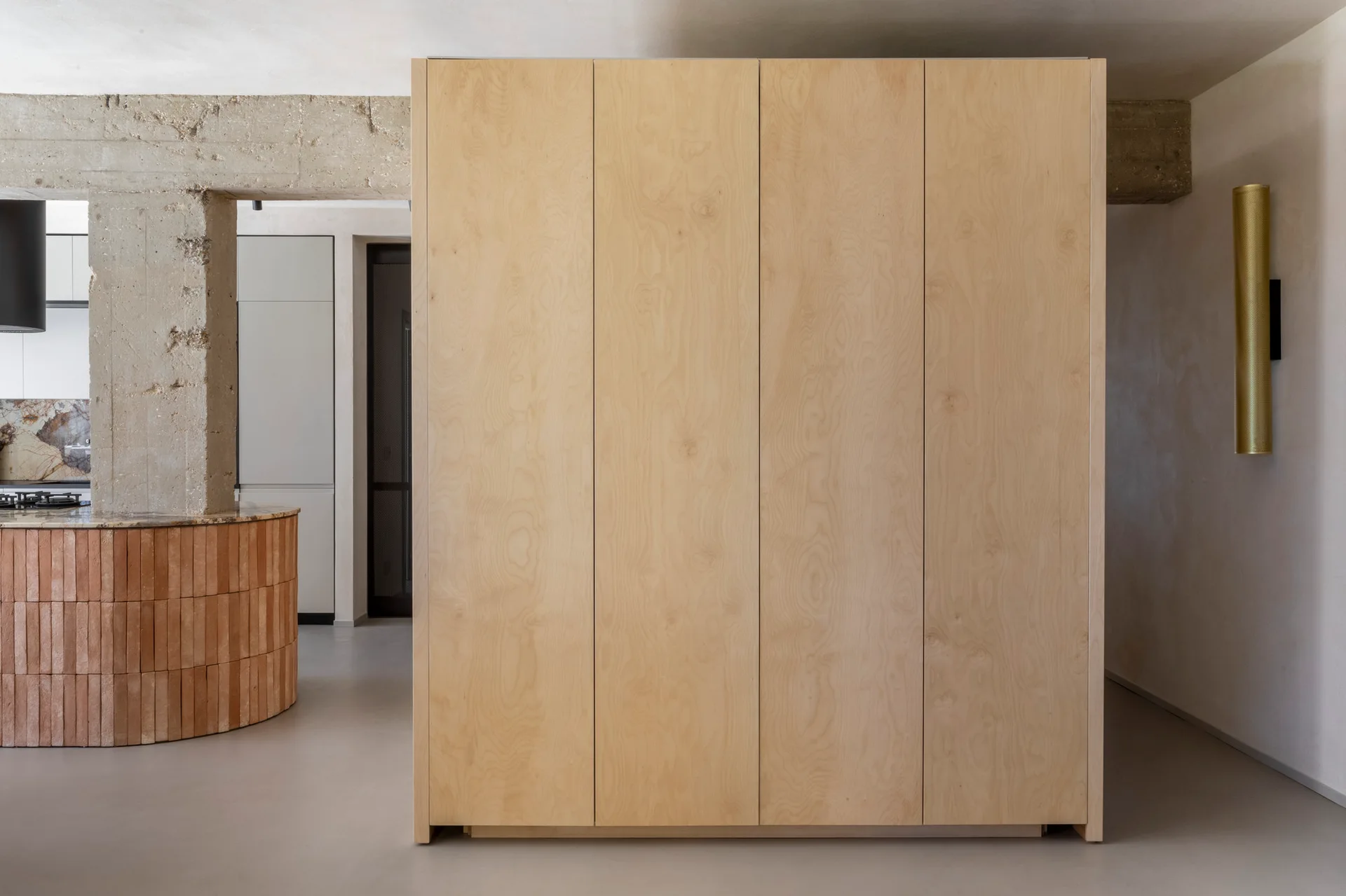 A minimalist room features a large light plywood cabinet standing beside an exposed concrete pillar and a curved brick kitchen island in the background.