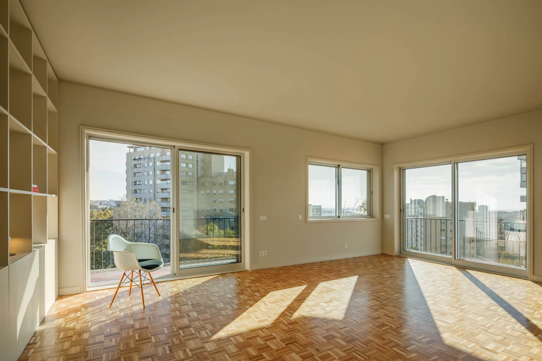 An empty apartment living room features a geometric parquet wood floor, beige walls with built-in shelving, and a single white chair near sliding doors.