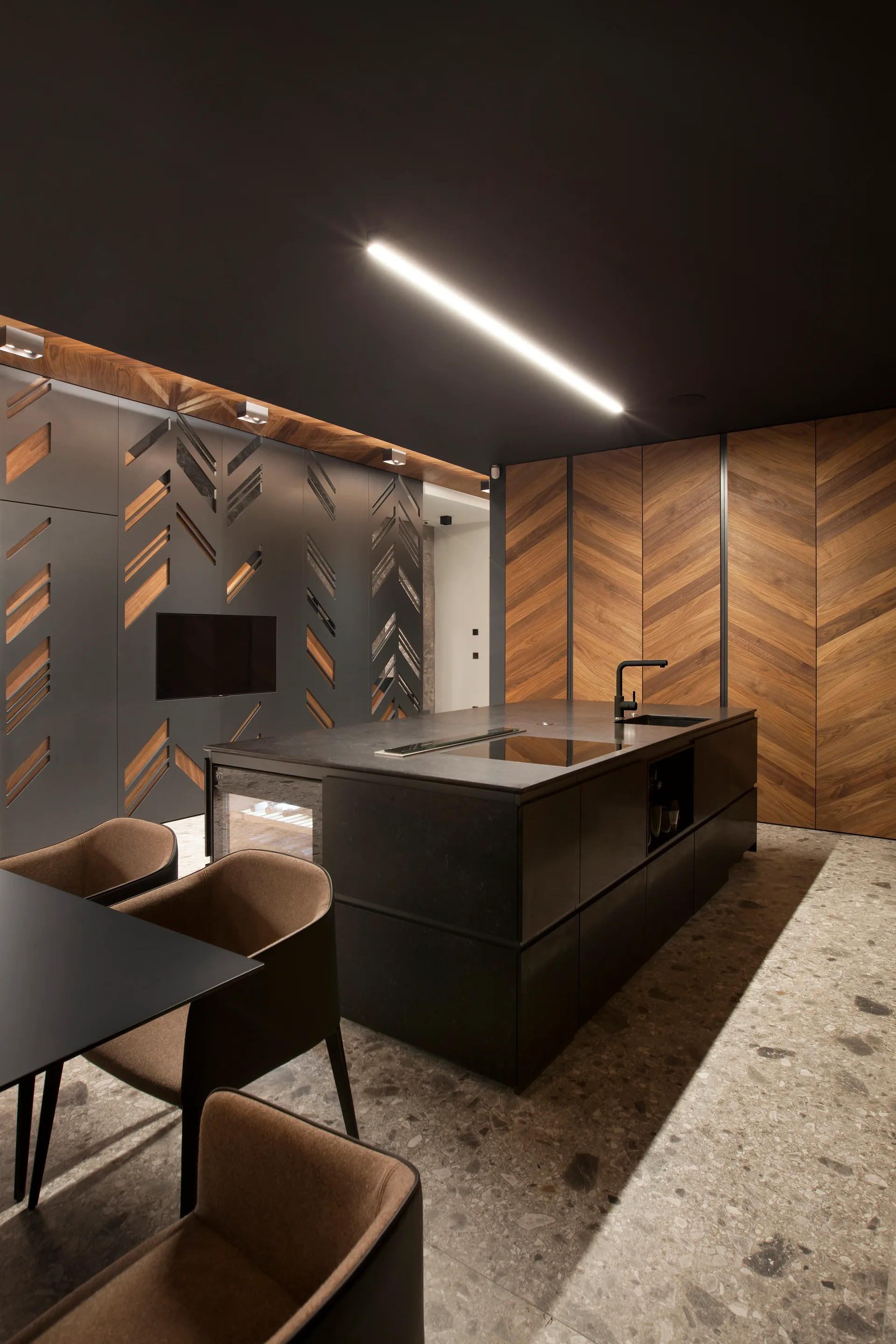A modern dark kitchen featuring a black island with a sink, chevron-patterned wood cabinet panels, a dark ceiling with linear lighting, and grey stone flooring.