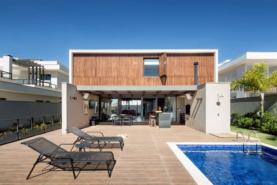 A modern patio features lounge chairs on a wood-look tile deck next to a blue swimming pool, with a wood-slatted facade on the house above.