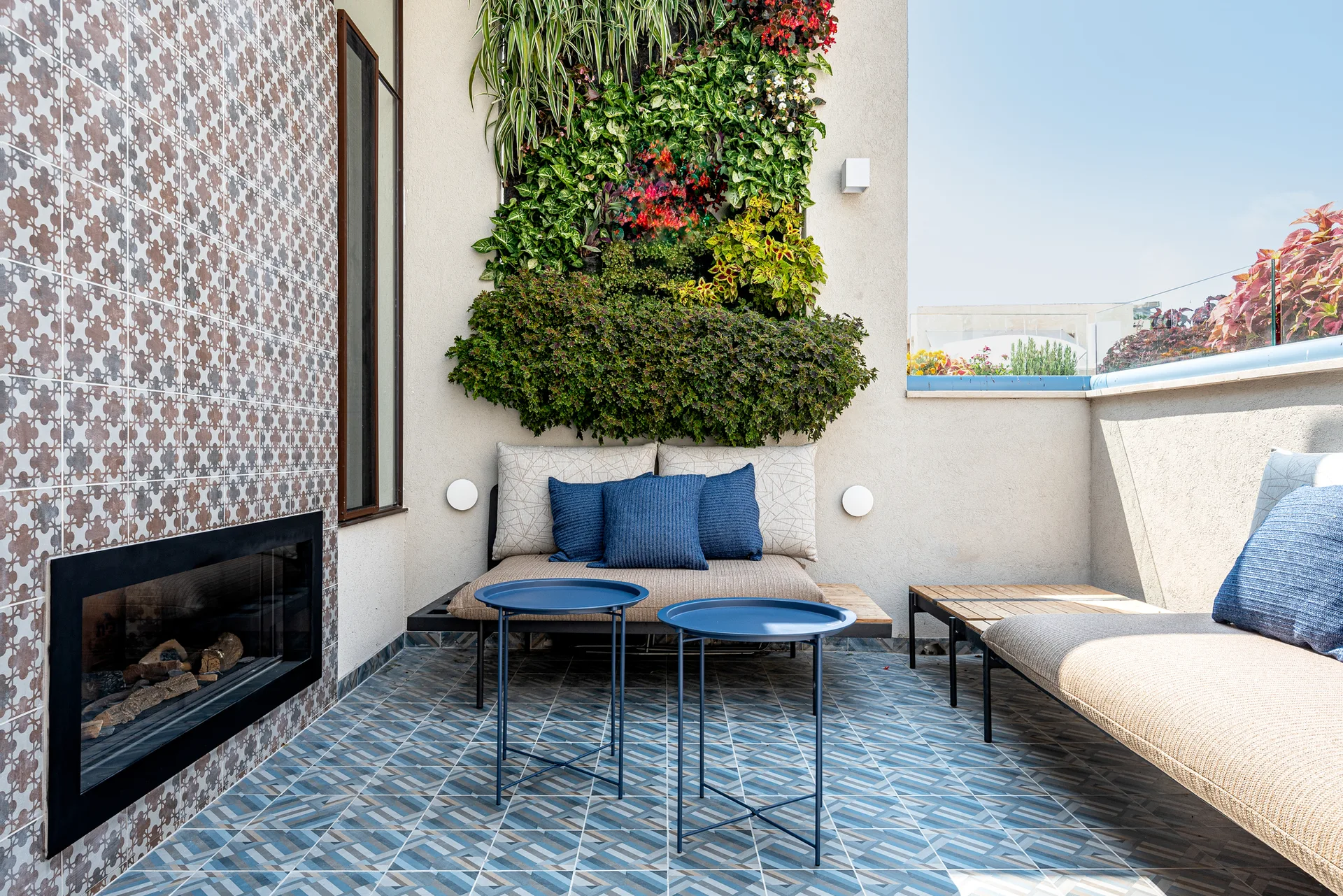 A modern outdoor terrace features blue geometric floor tiles, low-profile seating with blue pillows, a fireplace, and a lush vertical wall garden.