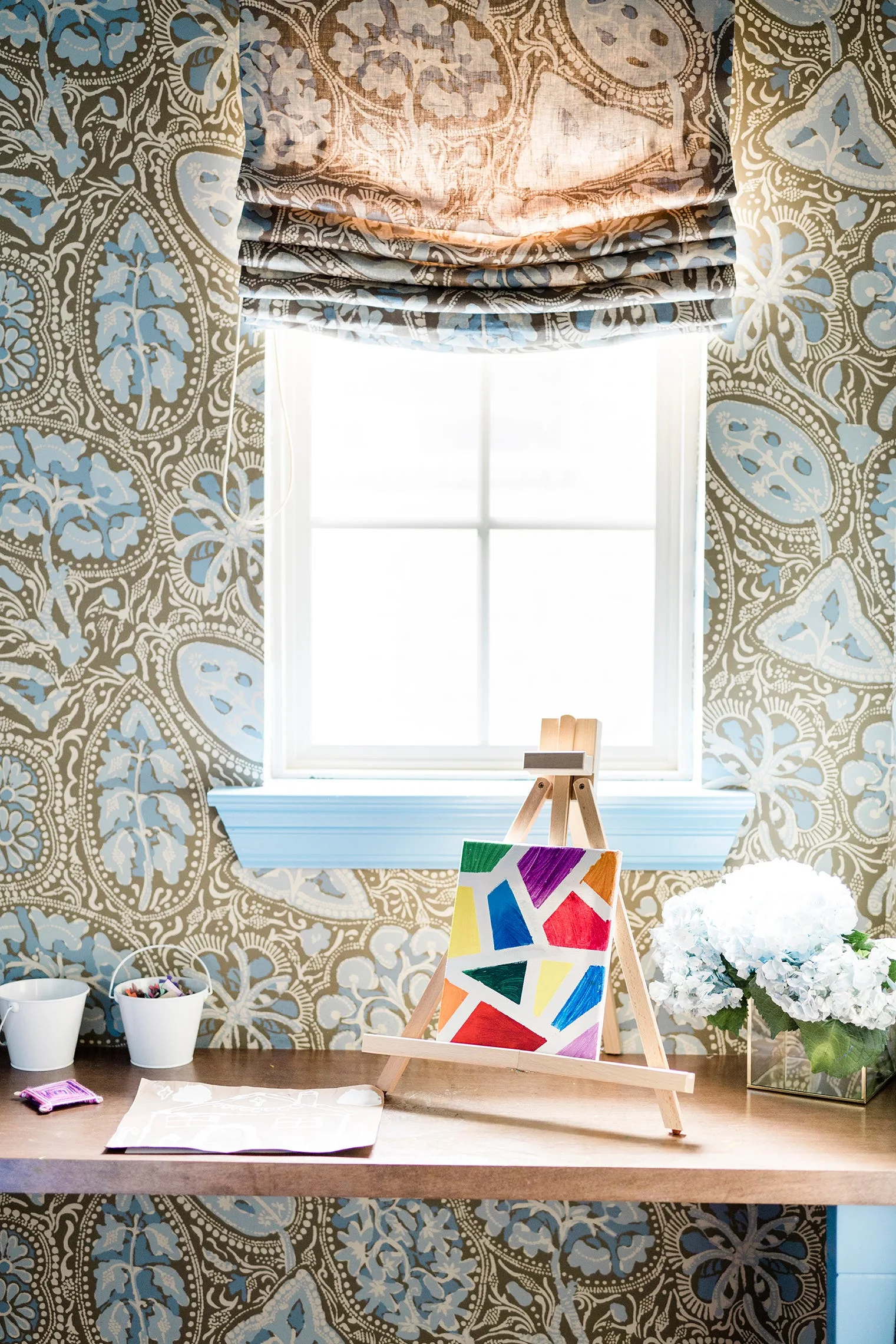 A small wooden easel with a colorful canvas sits on a desk in front of a window flanked by blue and brown patterned wallpaper.