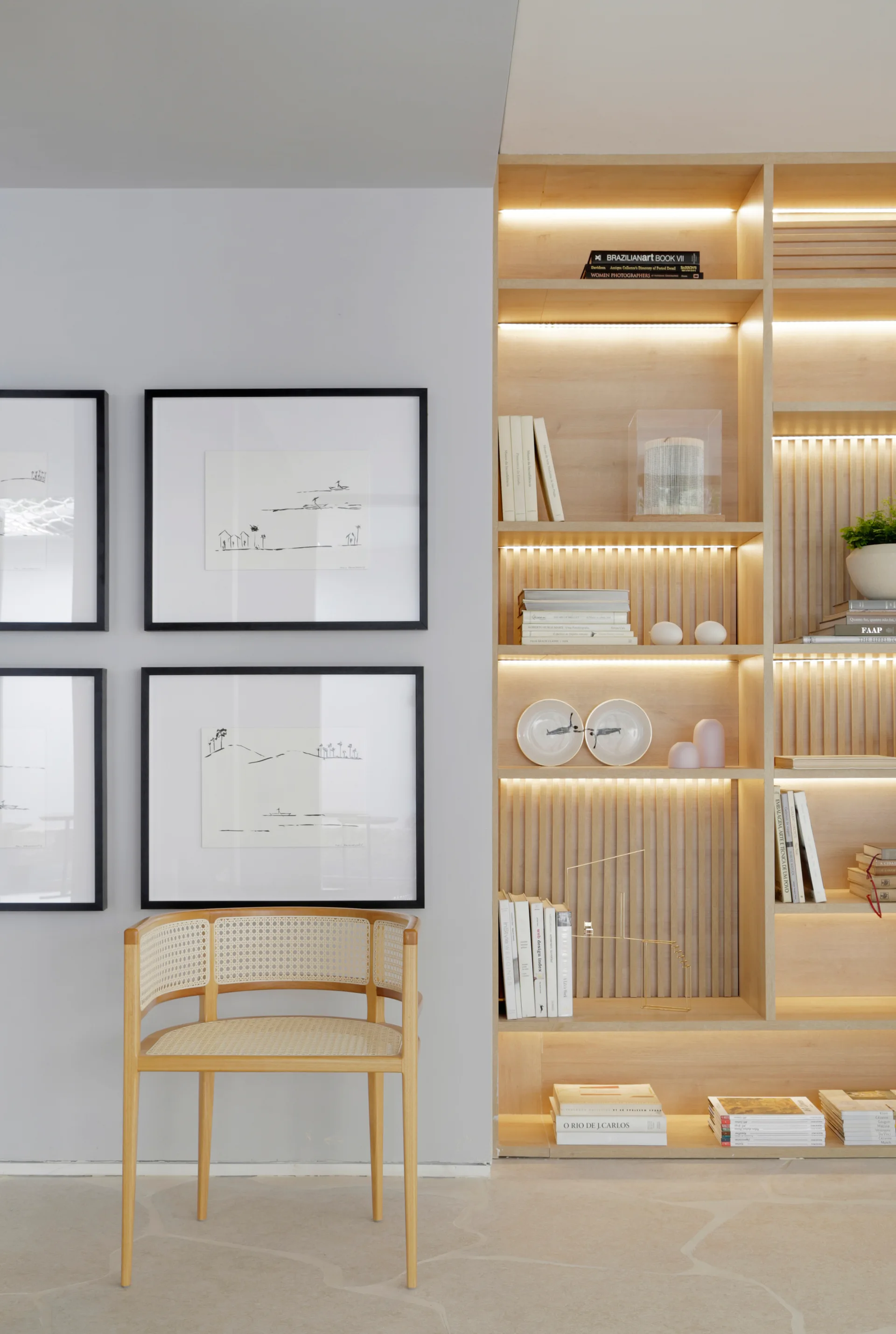 A light wood built-in shelving unit with warm LED lighting stands next to a grey wall featuring framed sketch art and a cane barrel chair.