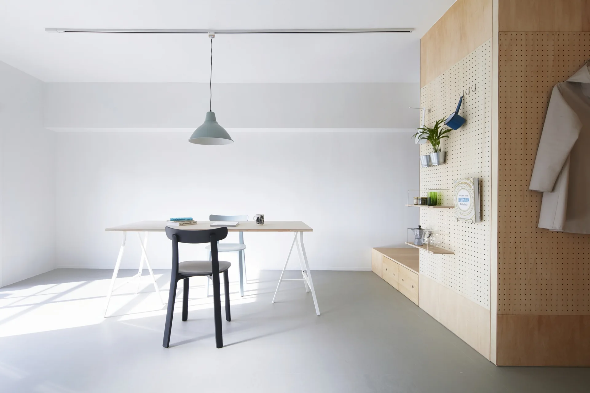 A minimalist dining room features a light wood trestle table with two chairs, a blue pendant light, and a large wooden pegboard wall storage unit.