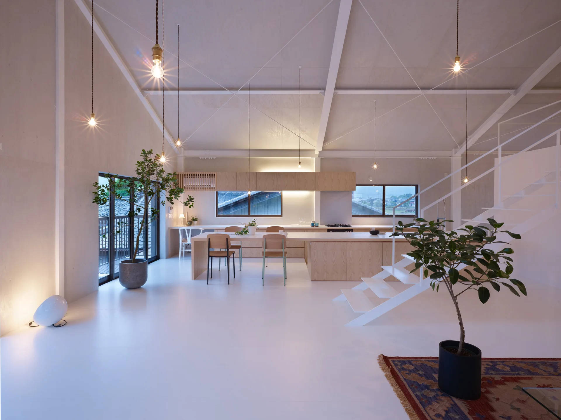 A minimalist loft kitchen with a light wood island, high vaulted ceiling, exposed beams, and a white steel open staircase.
