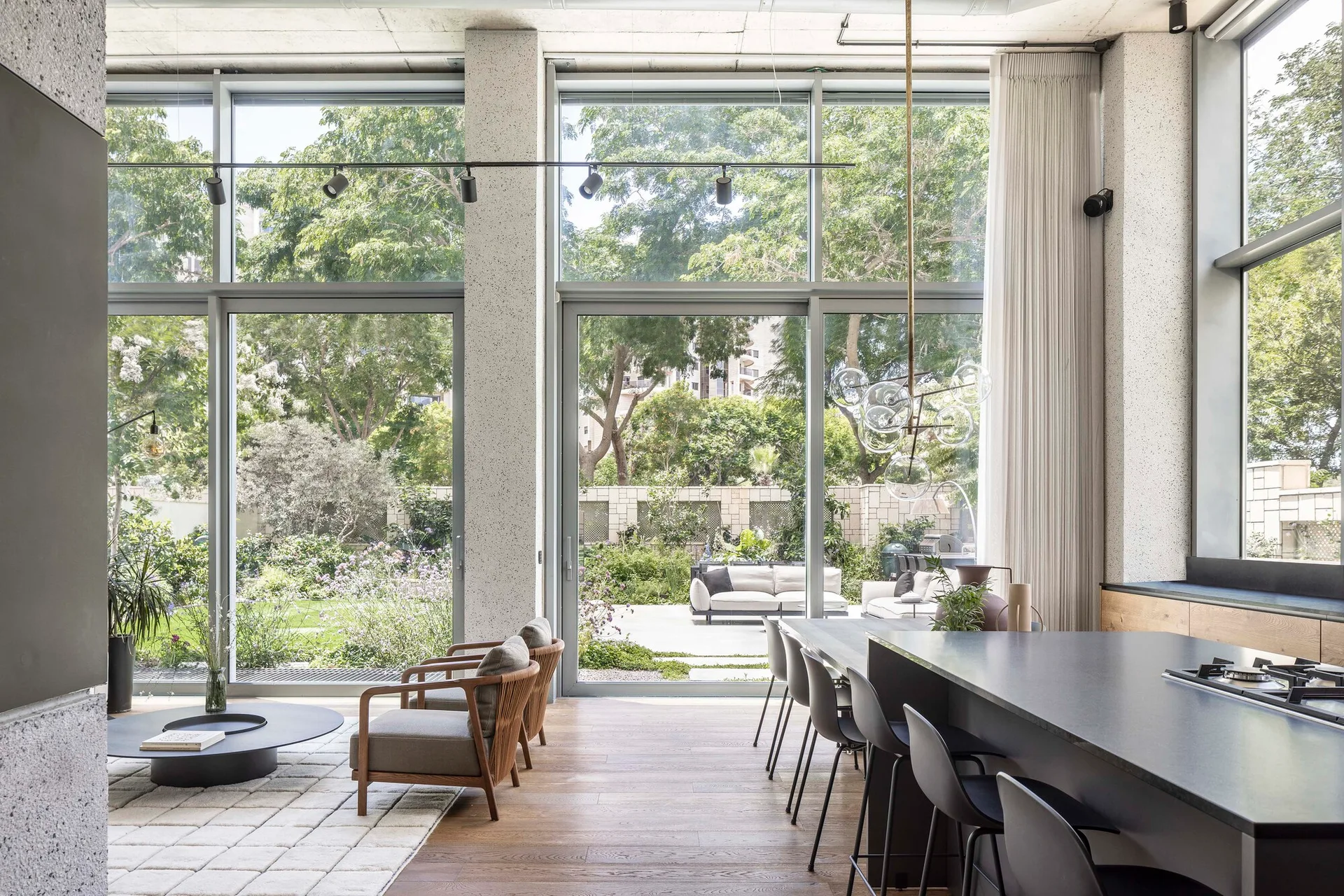 An open-plan living and kitchen area features tall glass walls facing a green garden, a dark kitchen island, and low wood-framed armchairs.