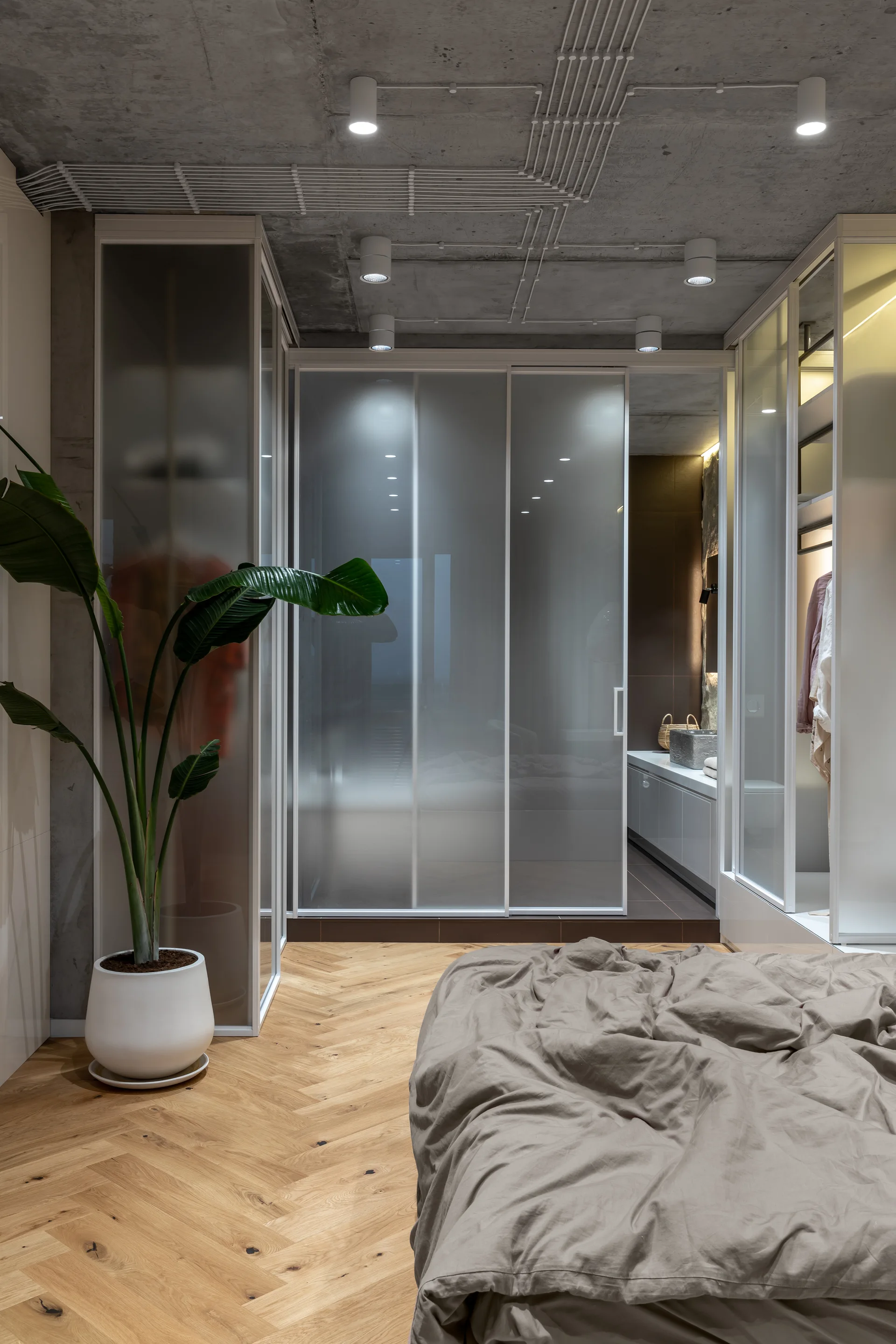 A modern bedroom with herringbone wood floors, a raw concrete ceiling, and large frosted glass sliding closet doors.