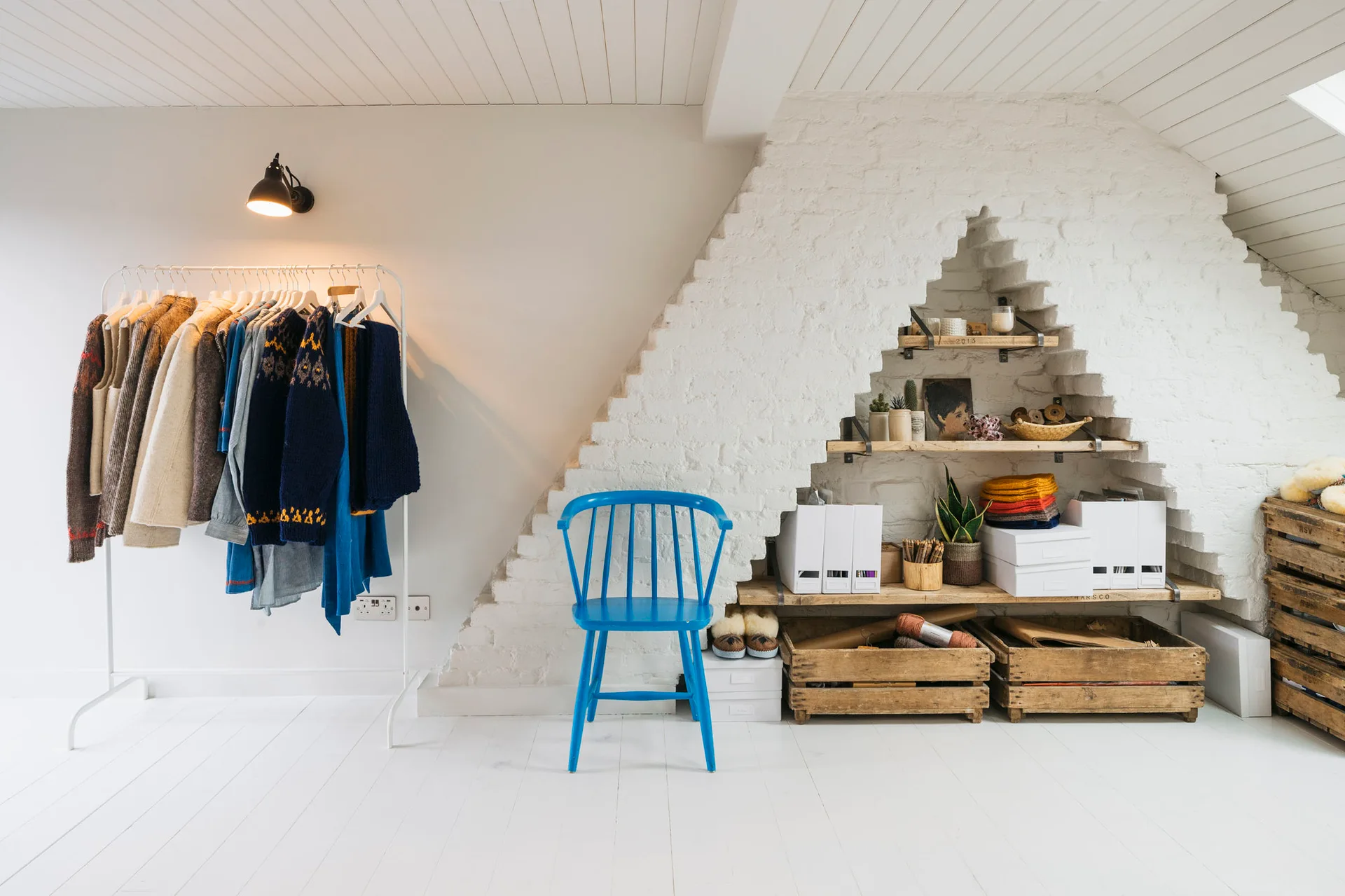 A bright attic dressing room features a white clothing rack, a bright blue wooden chair, and wood shelves built into a stepped white brick alcove.