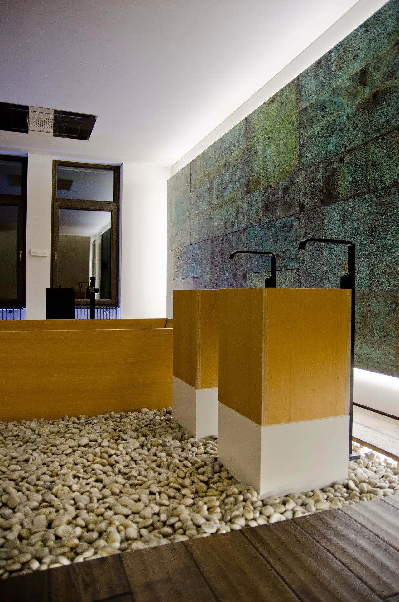 A modern spa bathroom features two wooden pedestal sinks with black faucets set against an oxidized copper tile wall and a river pebble floor.
