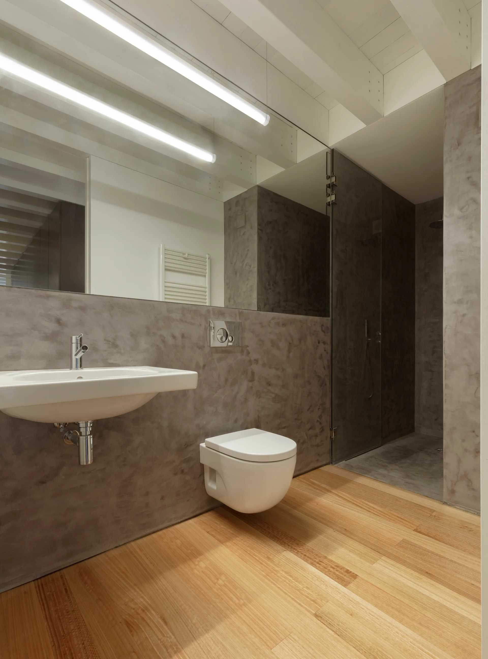 A minimalist bathroom featuring a wall-mounted sink and toilet against grey concrete-textured walls, with light wood flooring and a glass shower.