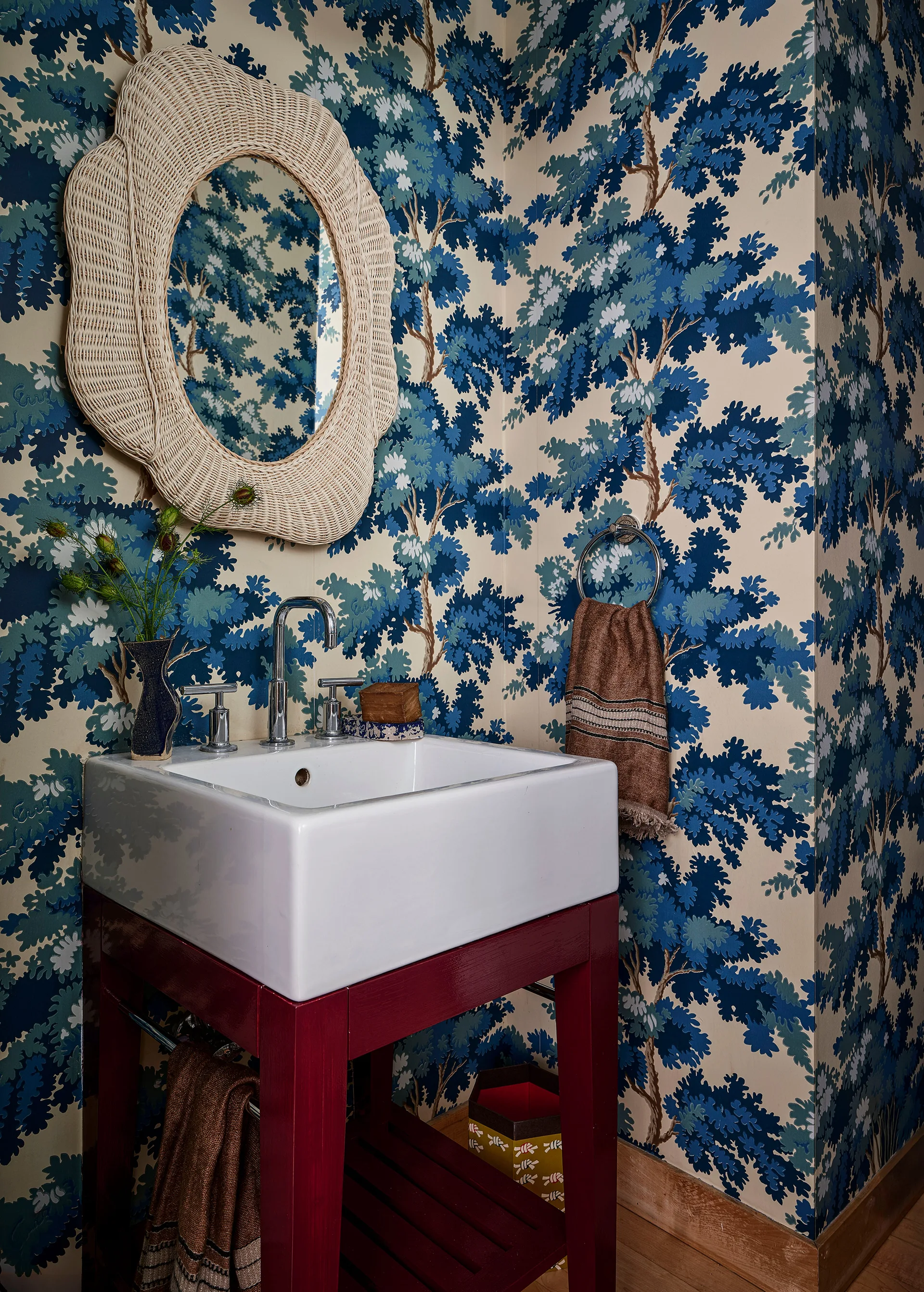 A powder room features blue and cream tree-patterned wallpaper, a dark red wood vanity with a white ceramic sink, and a scalloped rattan mirror.