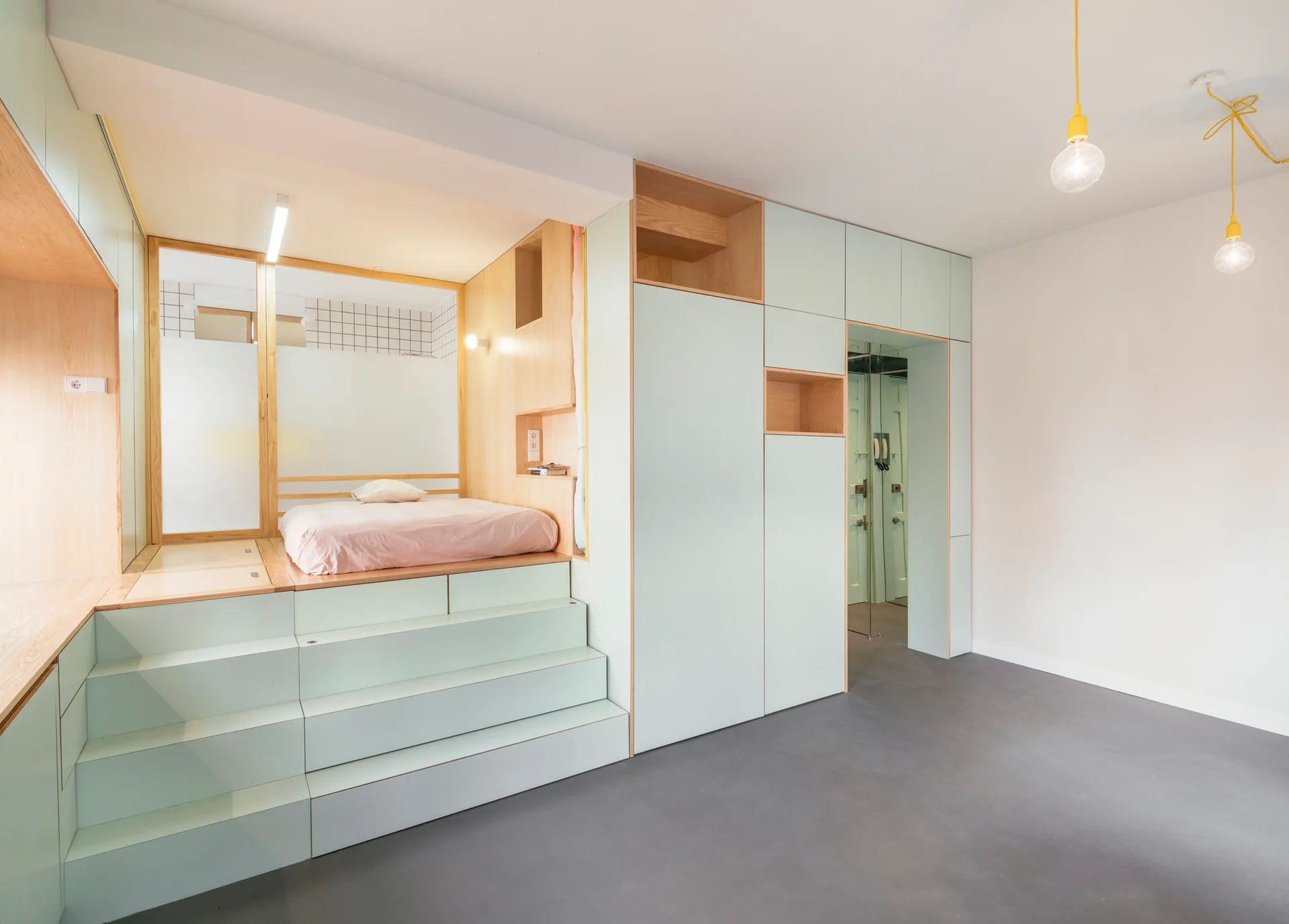 An elevated platform bed with pink bedding sits atop mint green storage stairs next to built-in light wood shelves and cabinetry.