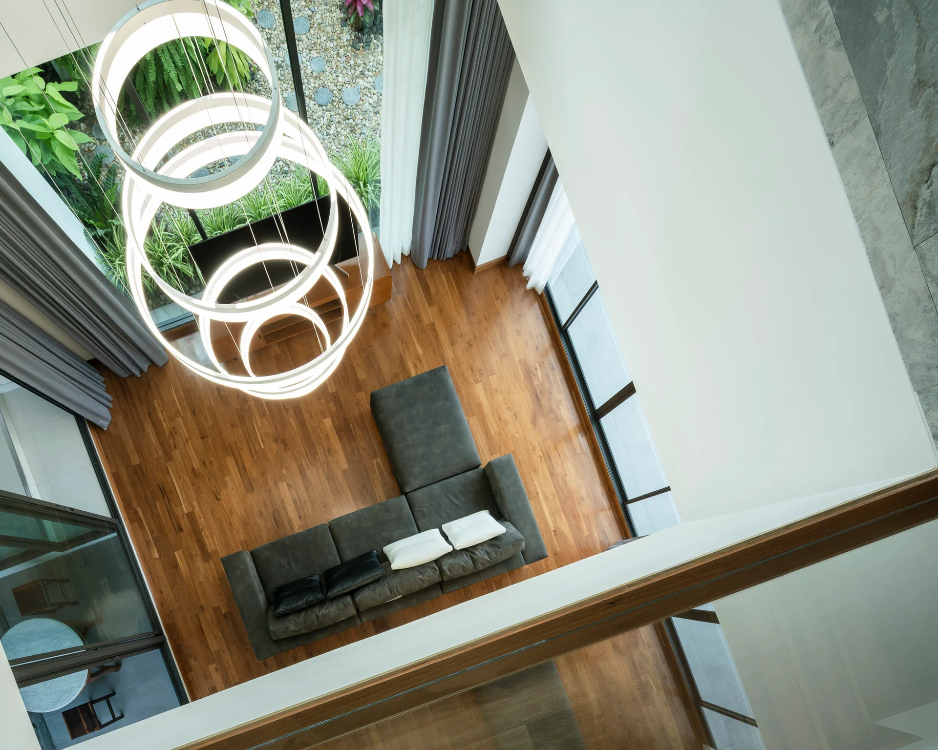 A high-angle view of a modern living room featuring a dark grey sectional sofa, warm wood flooring, and a large multi-ring LED pendant light.