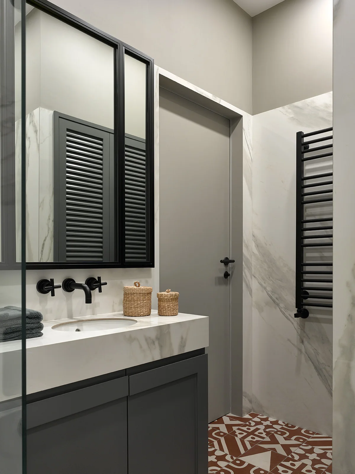A modern bathroom features a grey vanity with a white marble countertop, a wall-mounted black faucet, and patterned terracotta floor tiles.