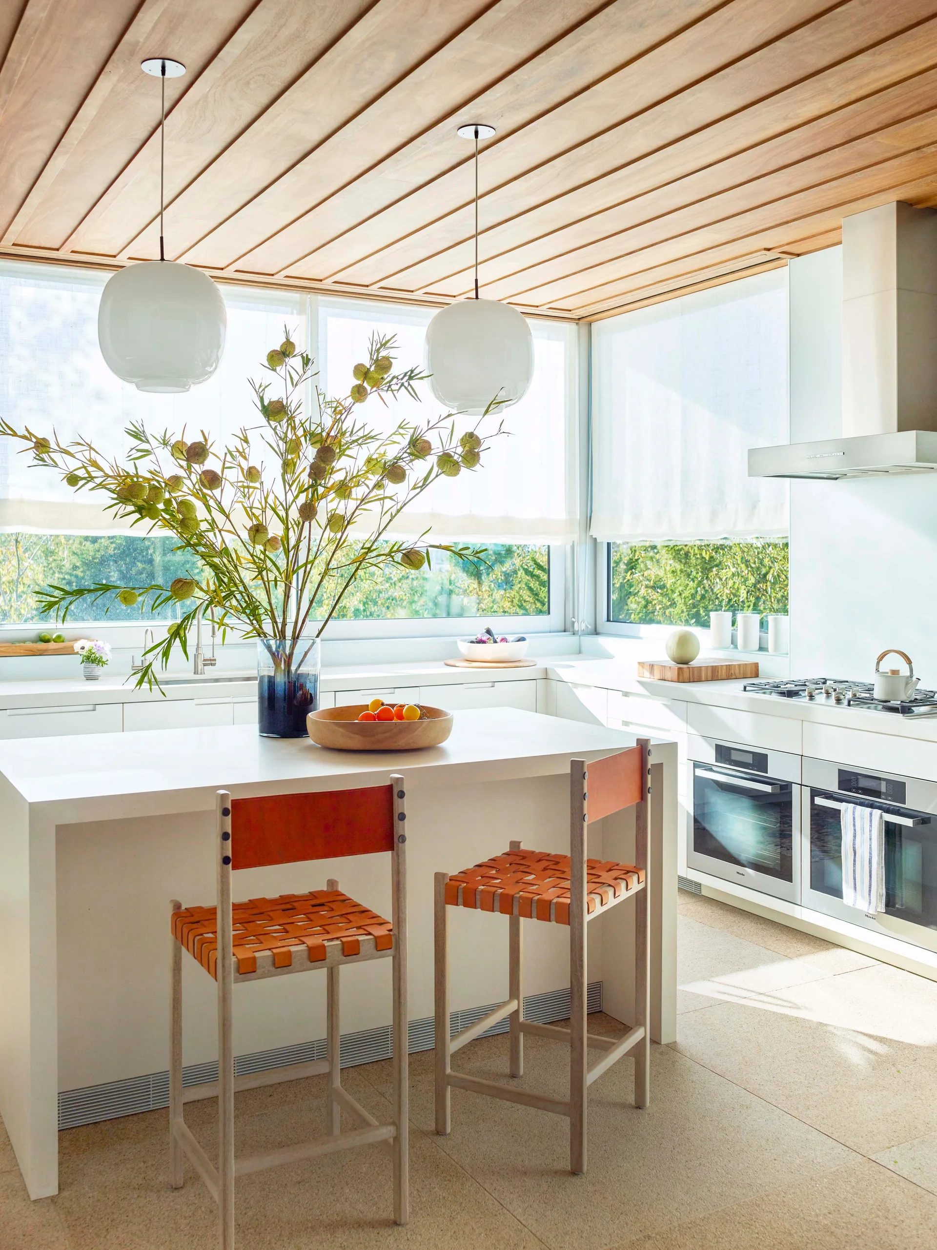 A modern white kitchen features a central island with woven orange leather stools beneath a natural wood plank ceiling and globe pendant lights.