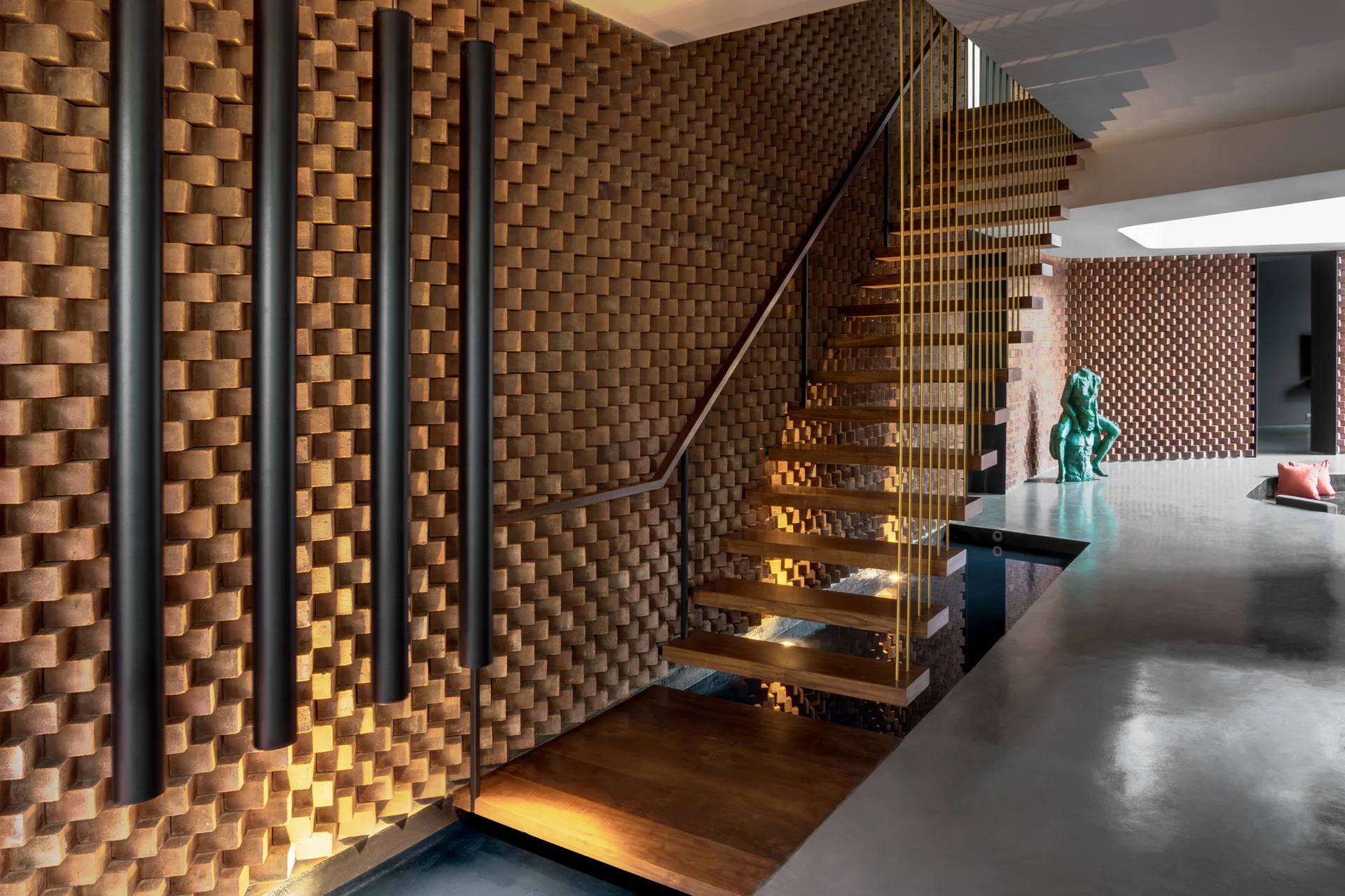 A floating wooden staircase with thin metal railings rises beside a textured brick wall and a polished concrete floor with a reflective pool.