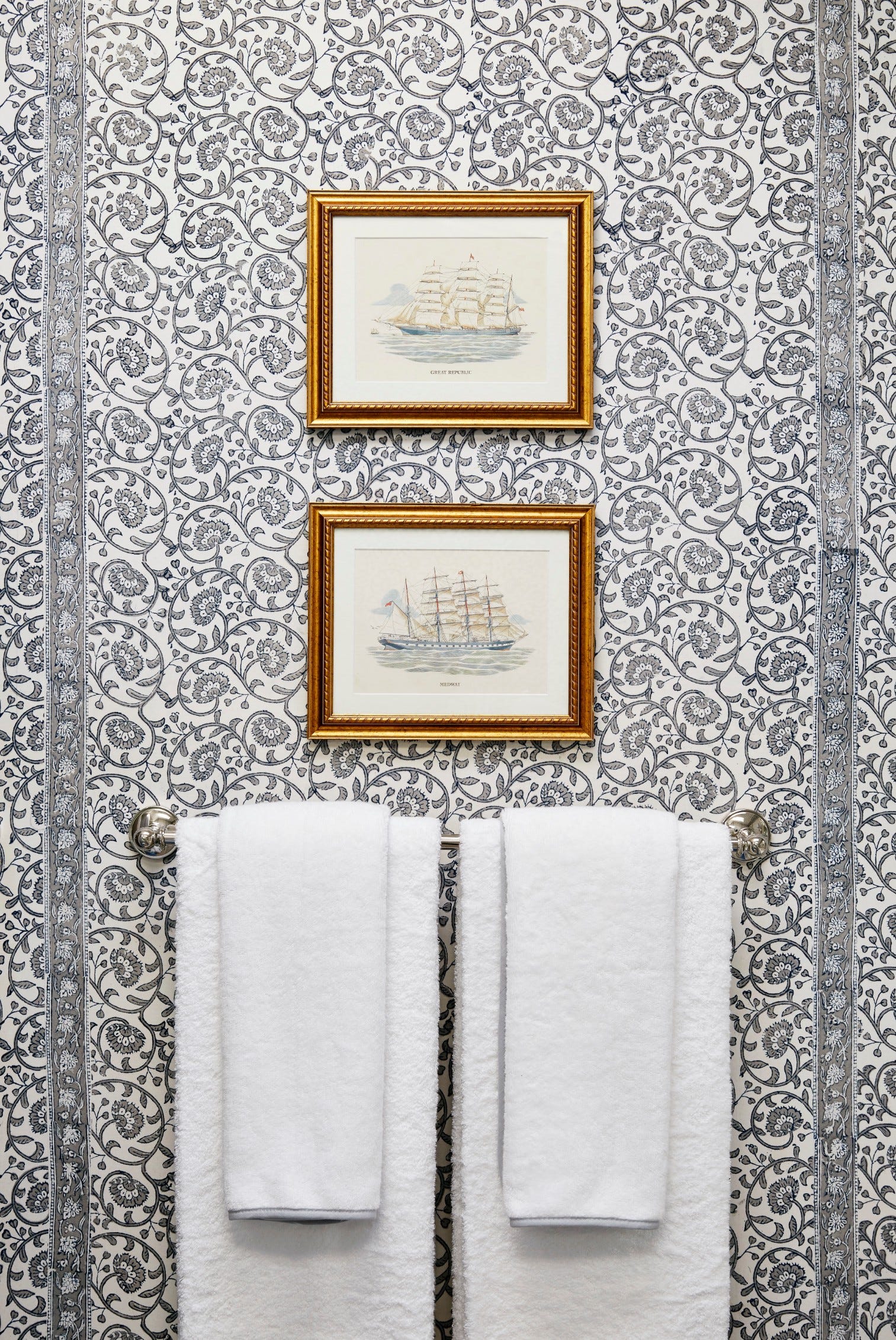 Two gold-framed nautical ship prints hang vertically on blue and white floral wallpaper above a chrome towel bar with white towels.