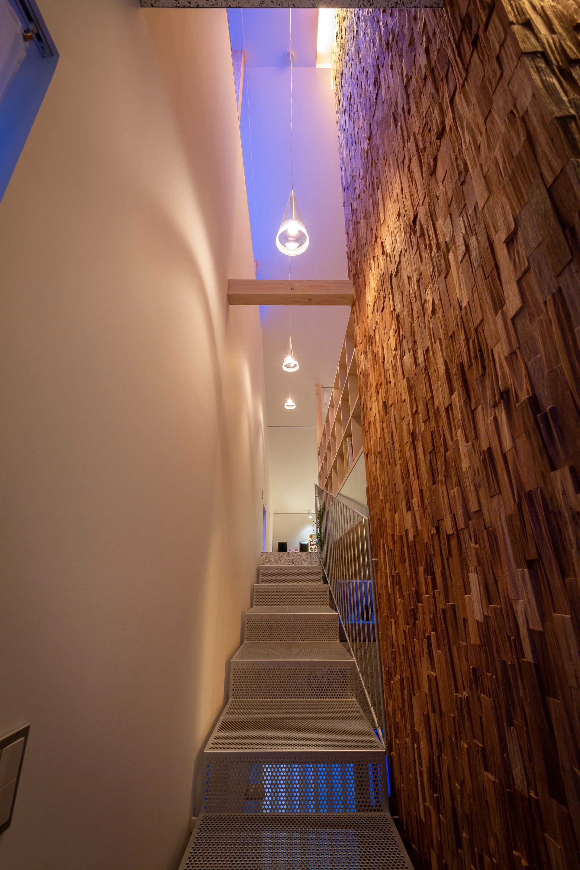 A modern staircase with perforated metal steps is flanked by a textured wood block feature wall and illuminated by pendant lights.