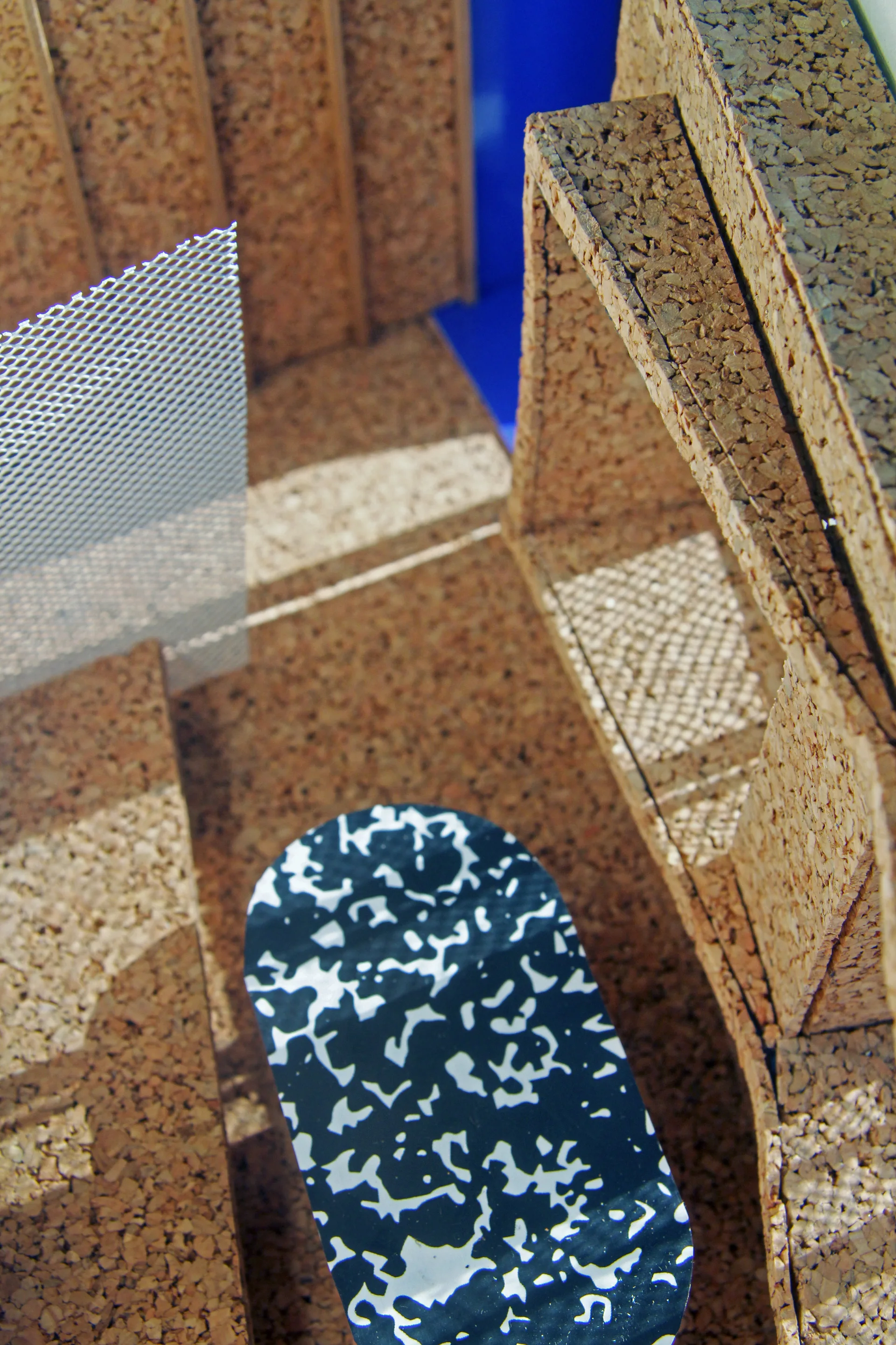 An architectural model displays textured cork panels, a metallic mesh partition, and a small black and white patterned oval table against a blue background.