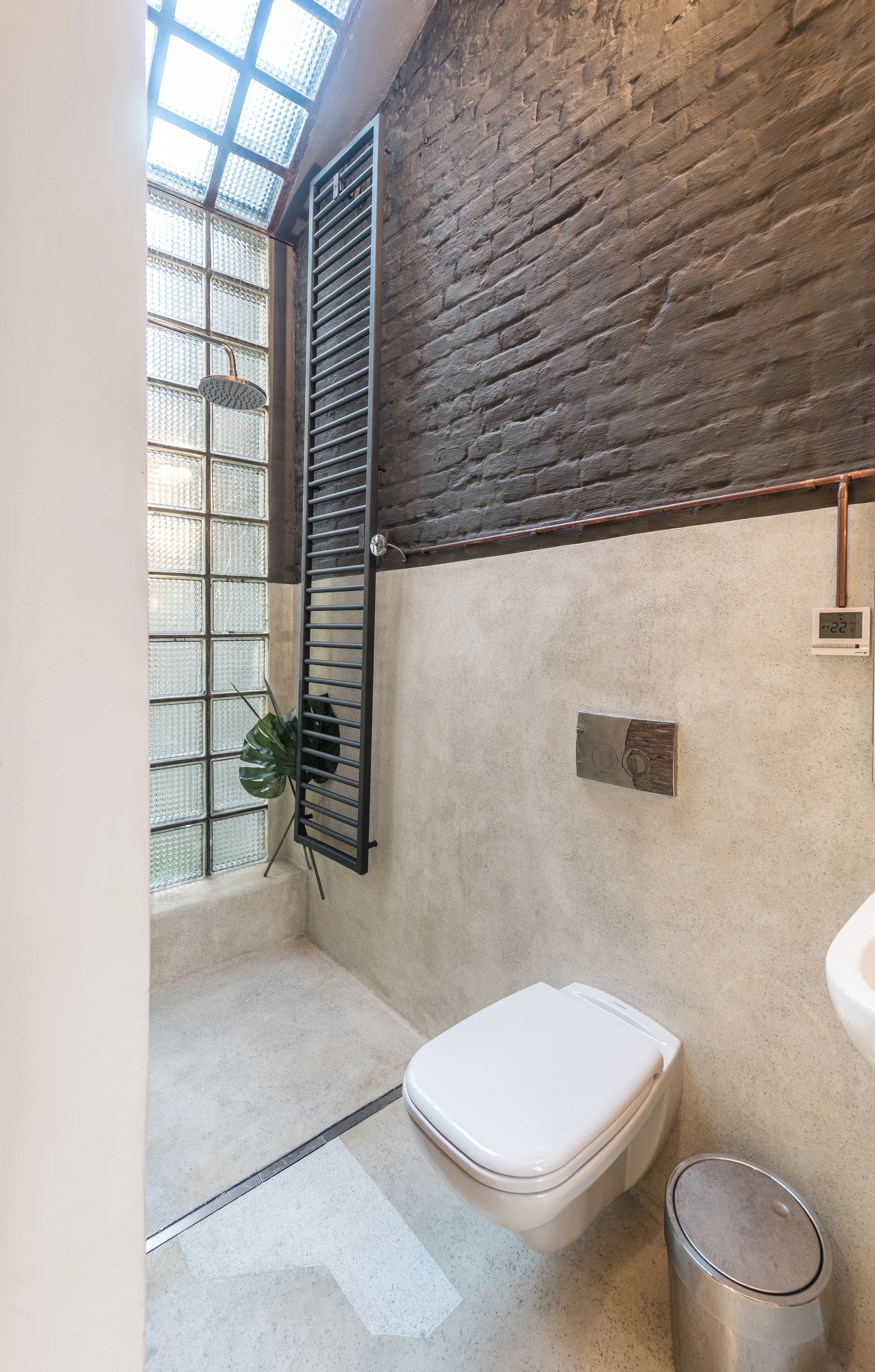 An industrial bathroom features a wall-mounted toilet next to a glass block shower wall, with a dark painted brick wall above smooth plaster.