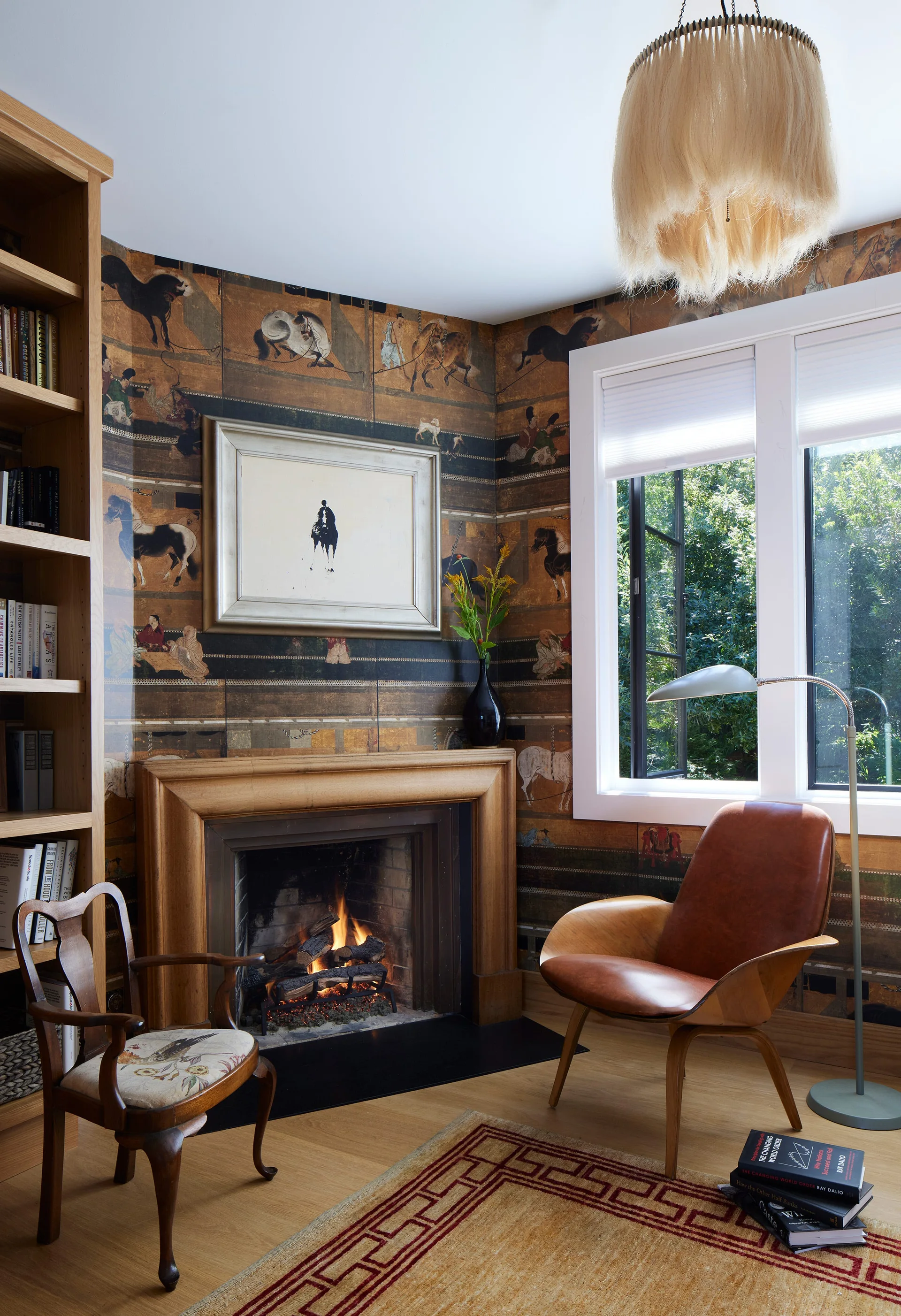 A cozy study corner features an active fireplace, patterned horse wallpaper, a brown leather lounge chair, and a light wood built-in bookcase.