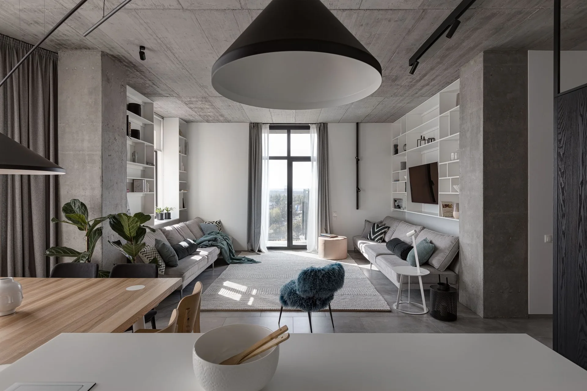A modern living room features two gray sofas facing each other on a textured rug beneath an exposed concrete ceiling and built-in white shelving.