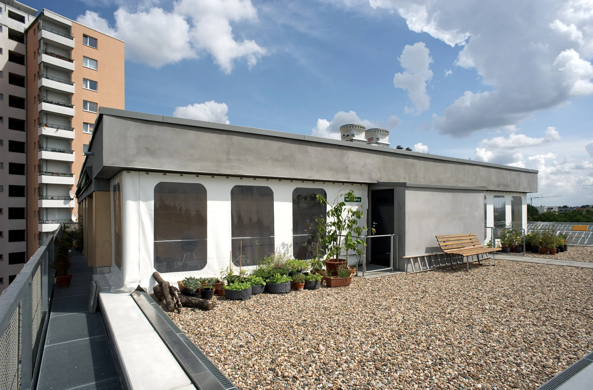 A rooftop terrace featuring a pebbled gravel floor, a wooden bench, potted plants, and a building facade with translucent panels under a partly cloudy sky.