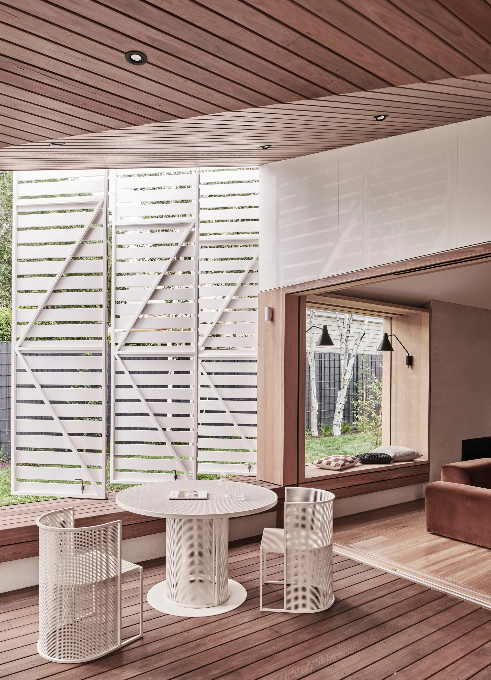 A covered wooden deck patio features a white circular metal table with matching mesh chairs next to tall, white louvered privacy screens.