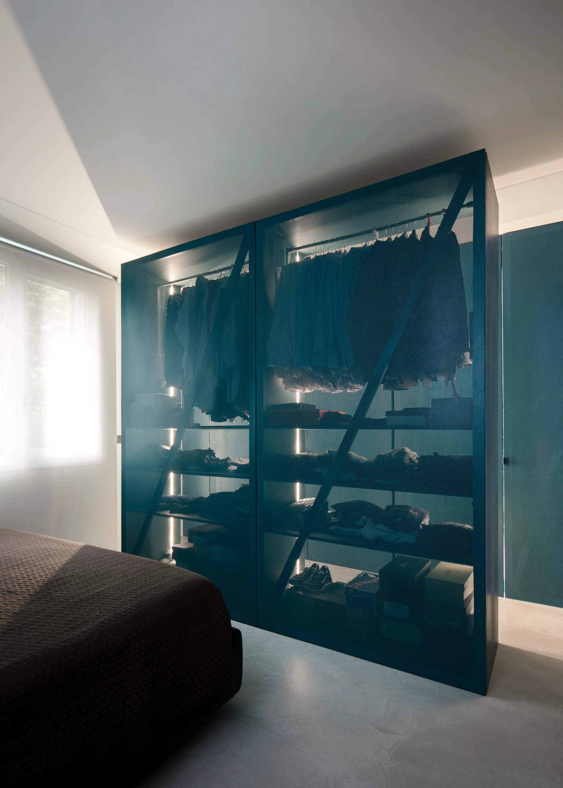 A dark blue perforated metal wardrobe with glowing vertical LED lights stands next to a brown bed on a smooth light gray floor.
