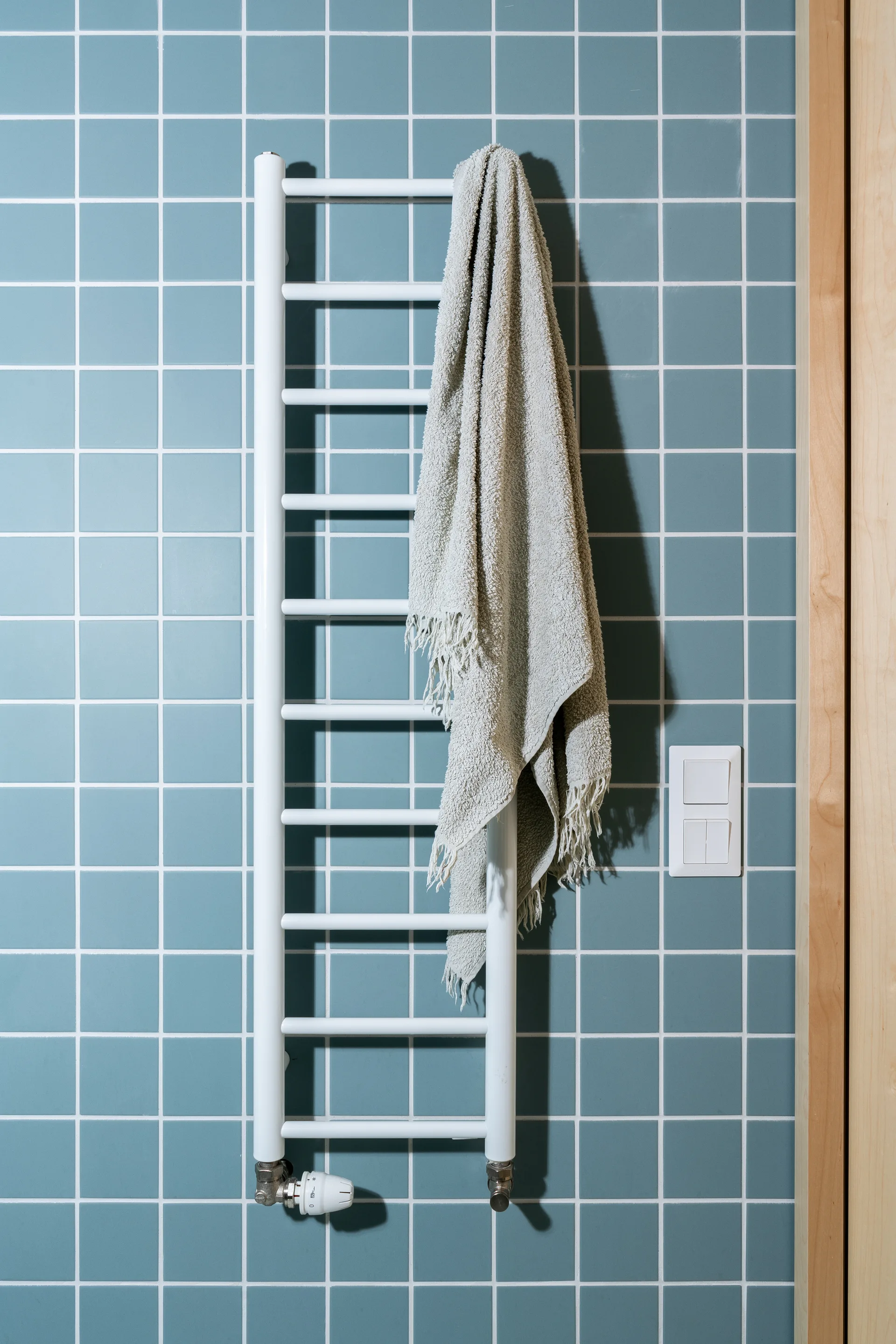A white ladder-style towel warmer with a grey fringed towel hangs on a blue square-tiled wall next to a double light switch.