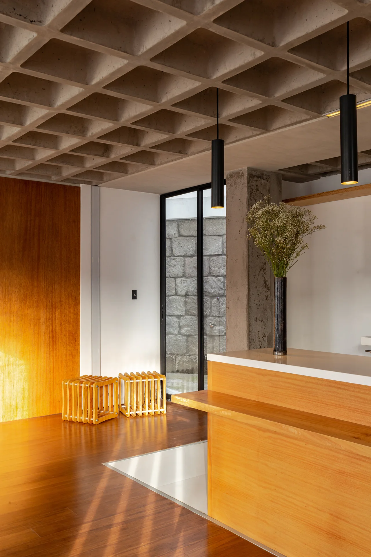 An interior space features a grey concrete waffle ceiling, warm wood flooring, two cylindrical black pendant lights, and a wooden kitchen island.
