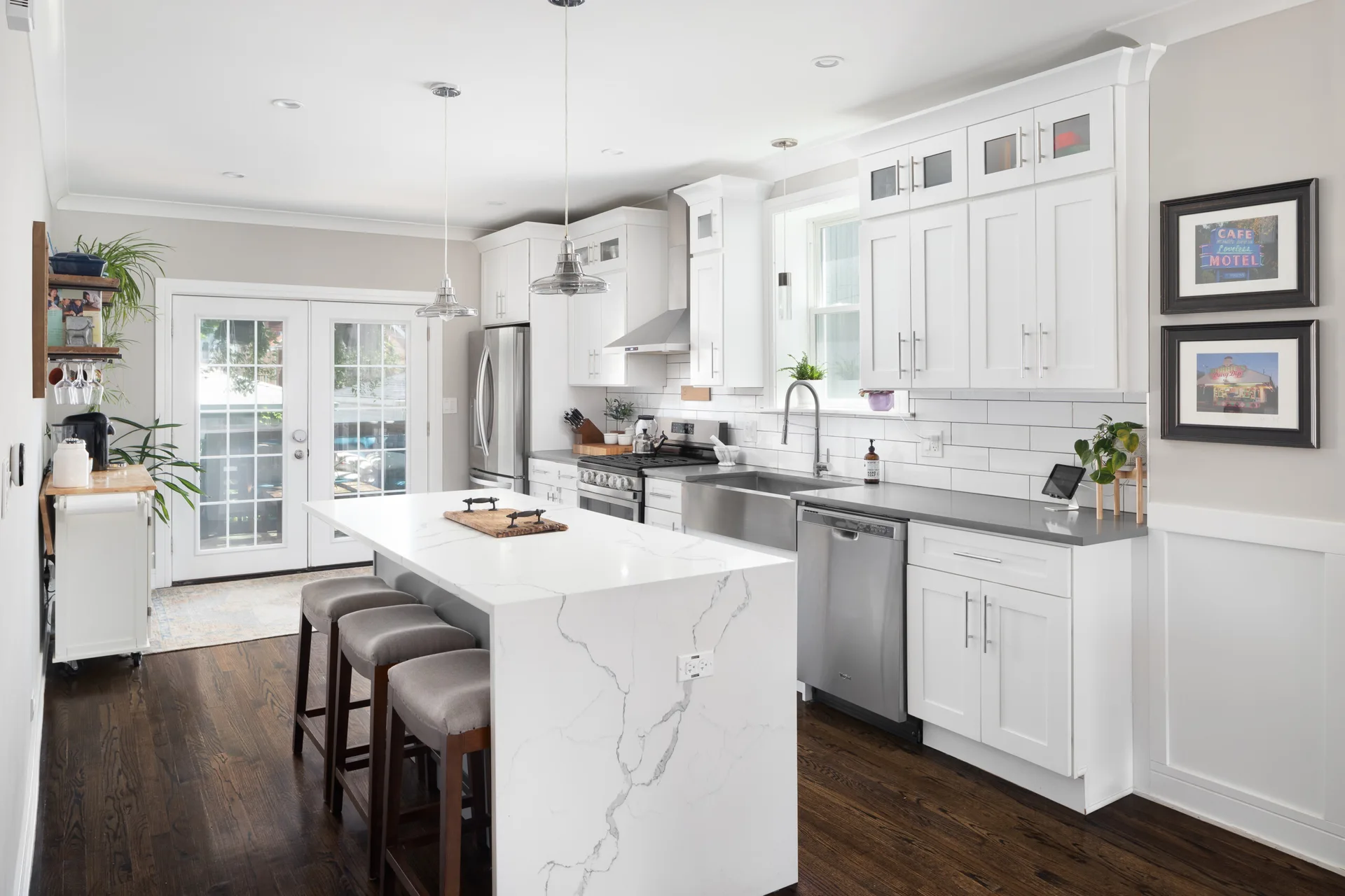 A bright kitchen featuring white shaker cabinets, a marble-veined waterfall island with three grey stools, and dark hardwood floors.