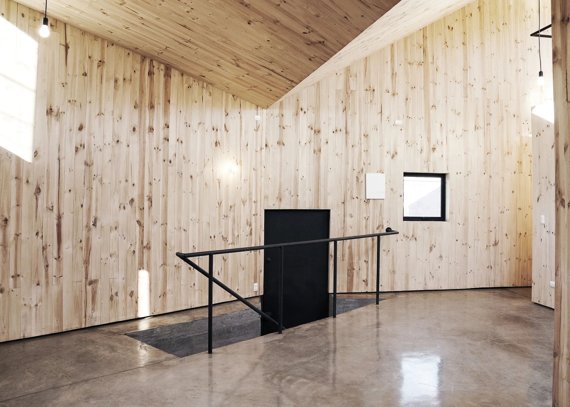 A minimalist staircase with a black metal handrail surrounded by light wood plank walls, an angled ceiling, and polished concrete flooring.