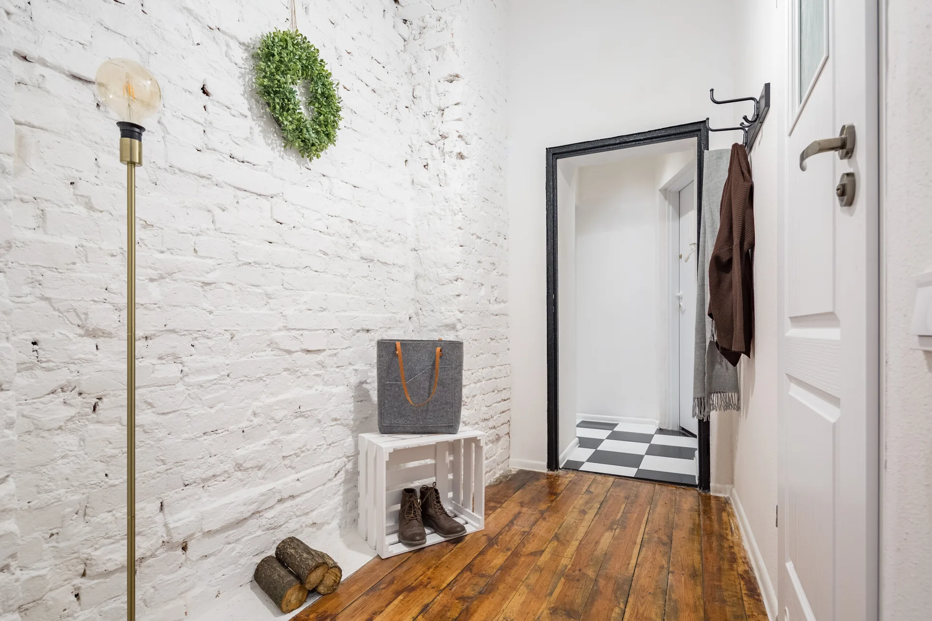 A rustic entryway features a white painted brick wall, warm hardwood floors, a white wooden storage crate, and a black wall-mounted coat rack.