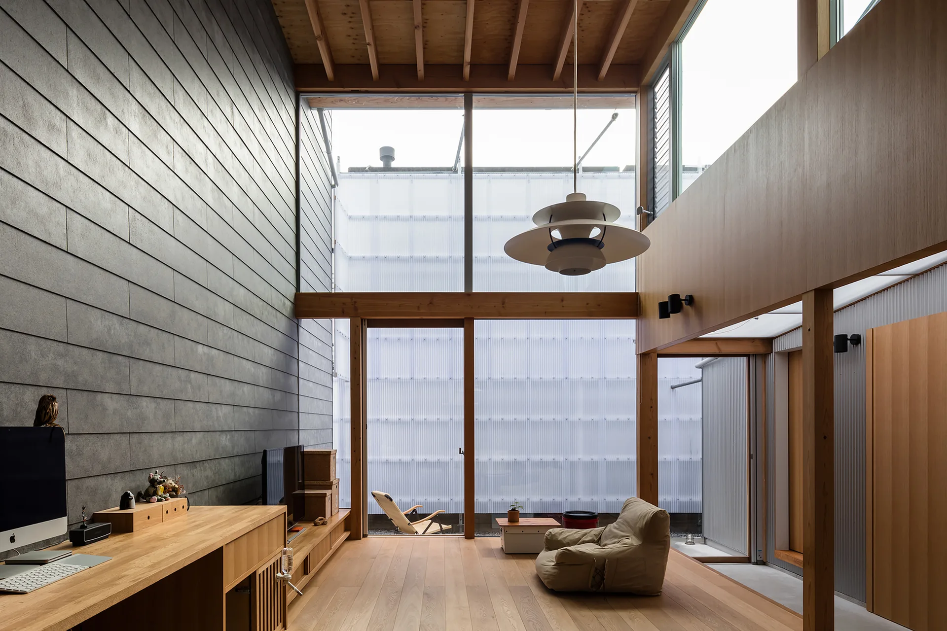 A modern living space with light wood floors, exposed ceiling beams, a grey paneled accent wall, a desk, and large sliding glass doors.