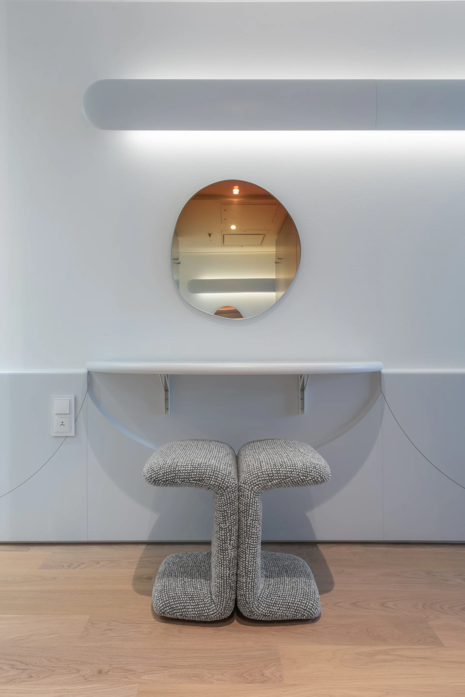 A minimalist white vanity features a wall-mounted console, a circular mirror, a linear wall light, and a sculptural grey upholstered stool on wood floors.