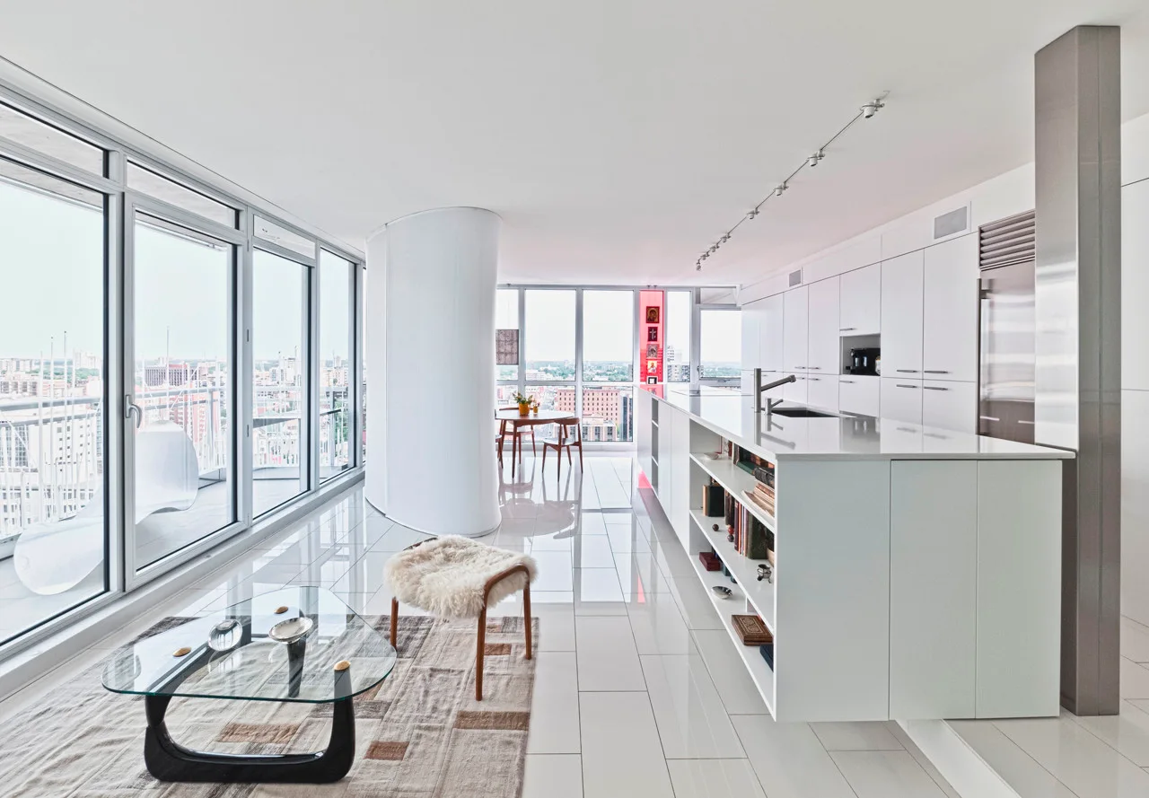 A modern open-plan kitchen and living space featuring floor-to-ceiling windows, a white island with open bookshelves, and a curved architectural partition.