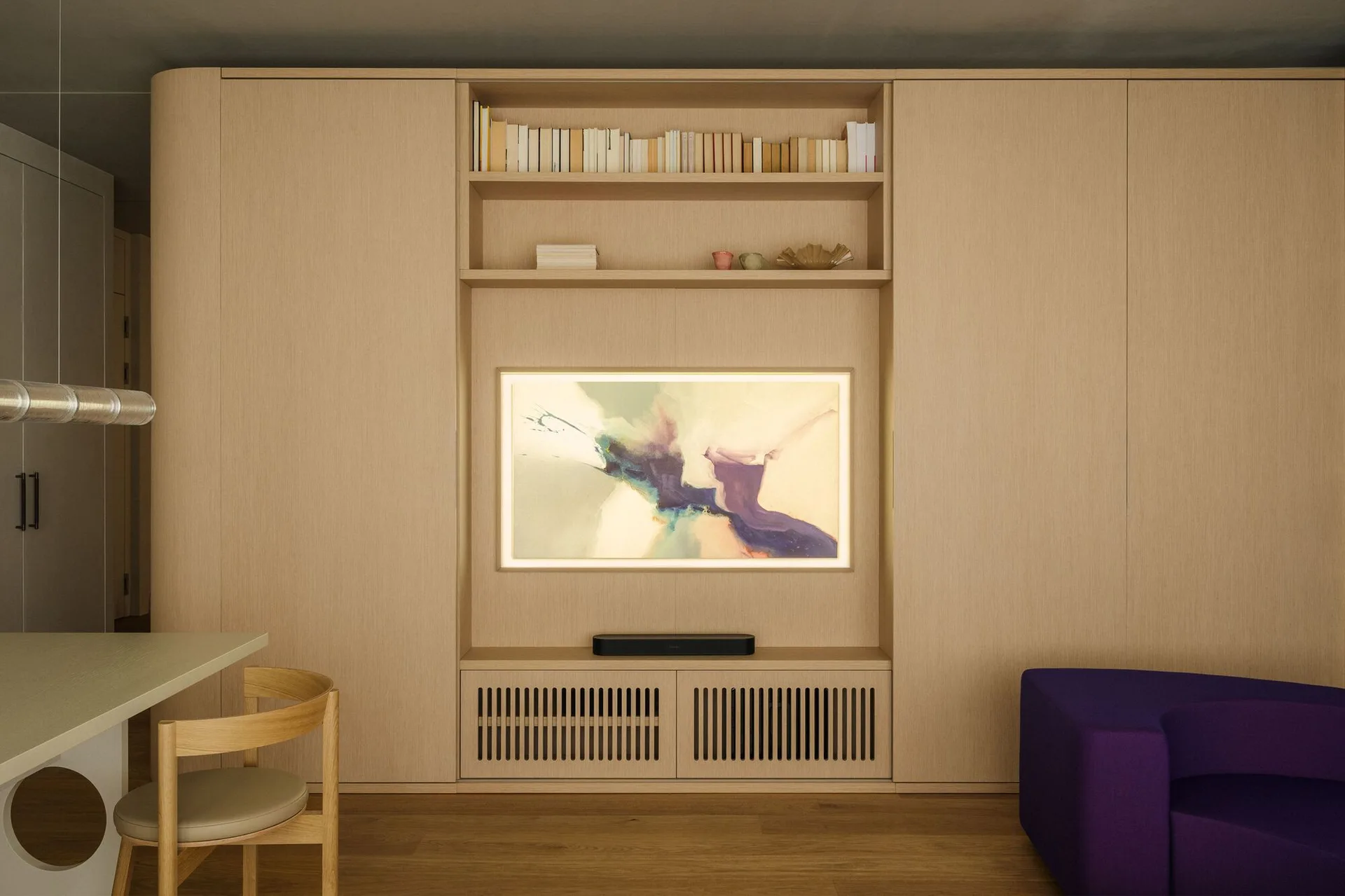 A minimalist living room features a light wood built-in cabinet wall with integrated shelving, a backlit screen showing digital art, and a purple sofa.