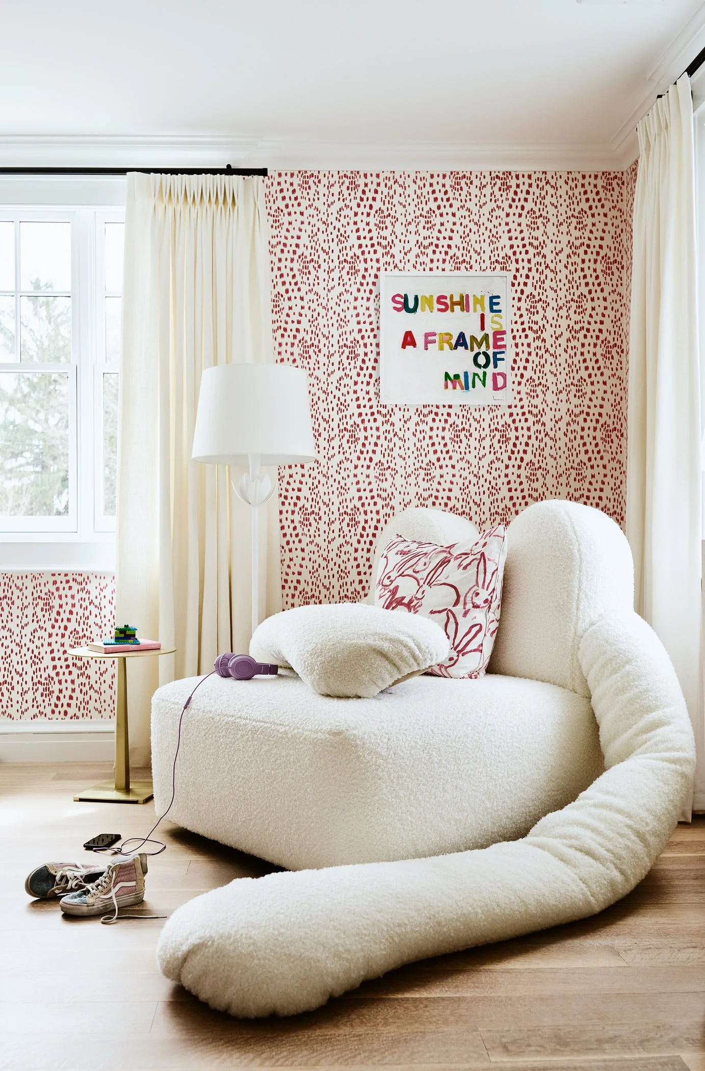 A whimsical kids room corner featuring an oversized cream boucle armchair with a trailing arm resting on wood floors beside red patterned wallpaper.