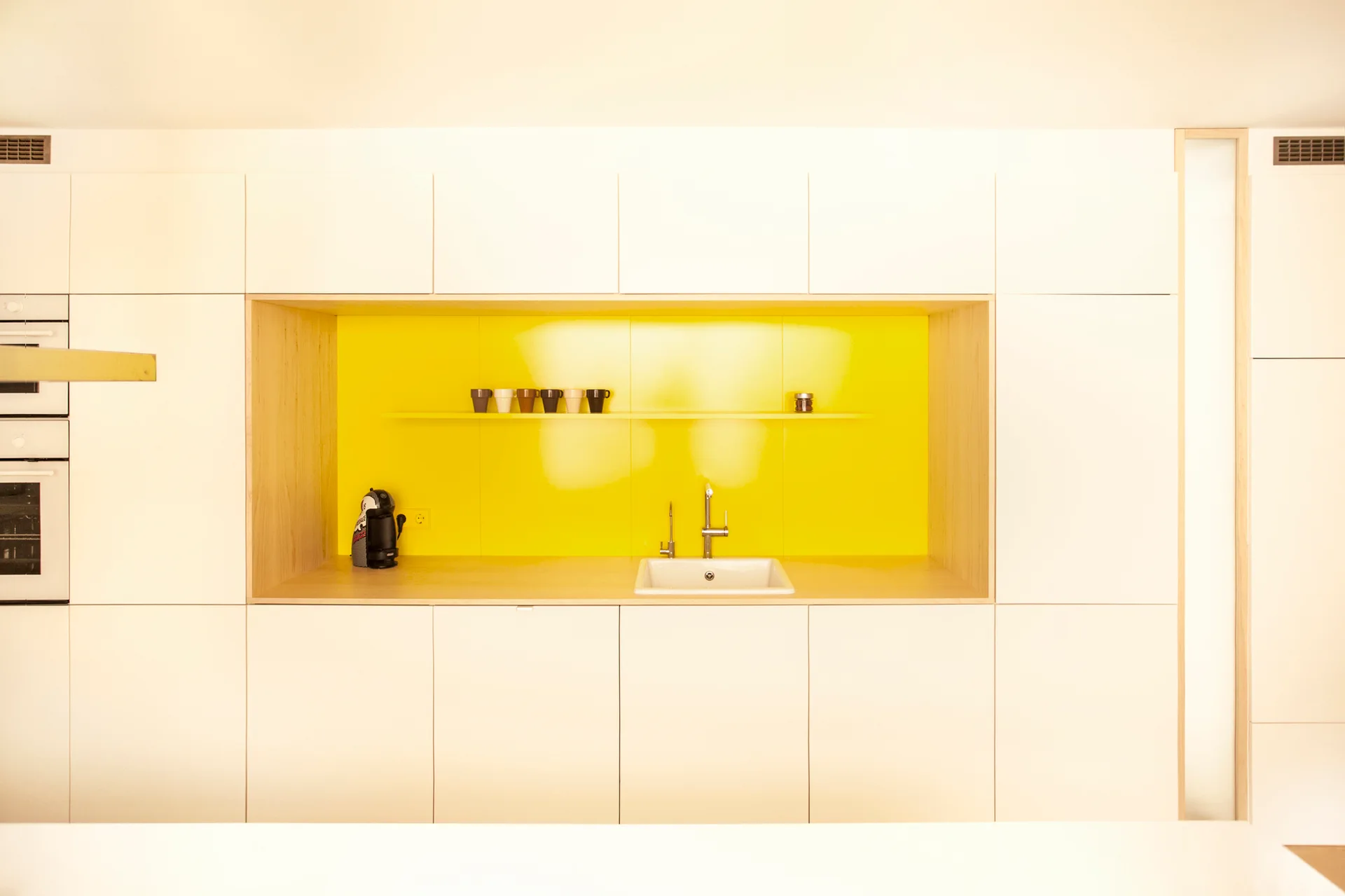 Sleek white cabinetry surrounds a recessed yellow niche with a light wood frame, housing a white undermount sink, a faucet, and a thin display shelf.