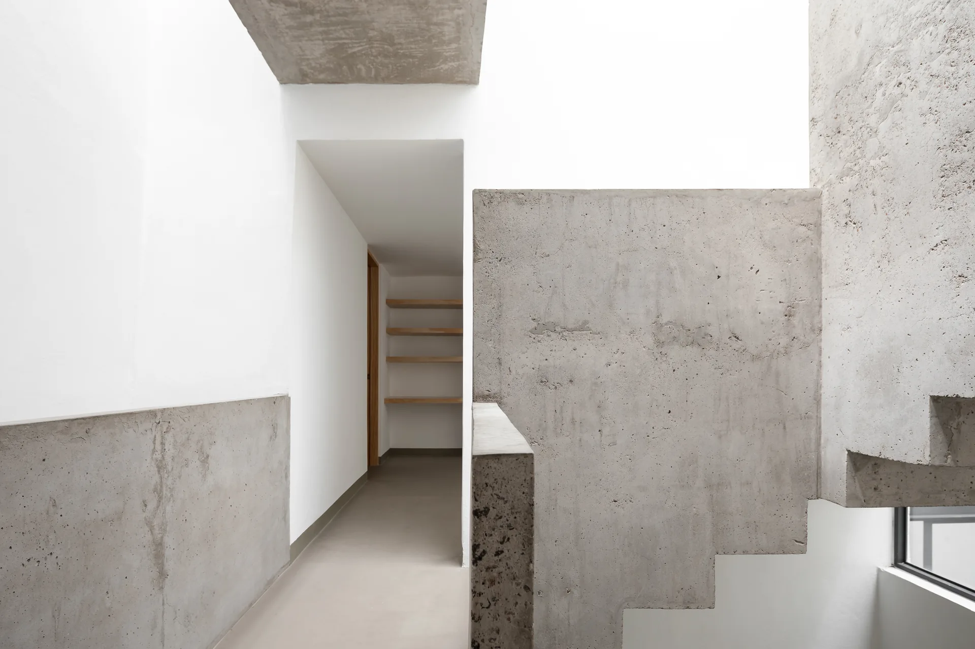 A minimalist hallway features a raw concrete staircase and walls contrasted against smooth white surfaces with recessed wooden shelves.