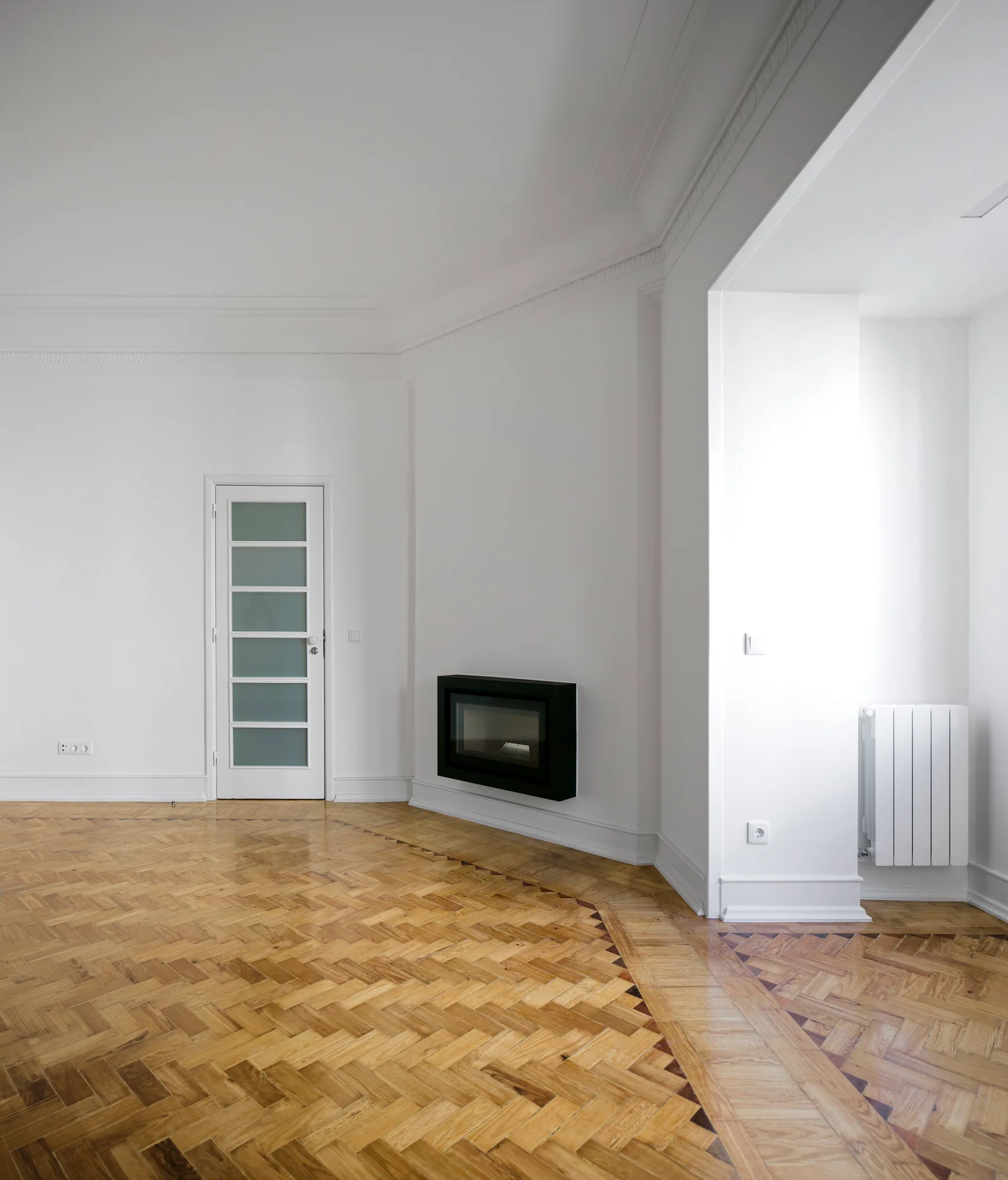 An empty white room with ornate crown molding, herringbone wood floors, a glass-panel door, and a black wall-mounted fireplace.