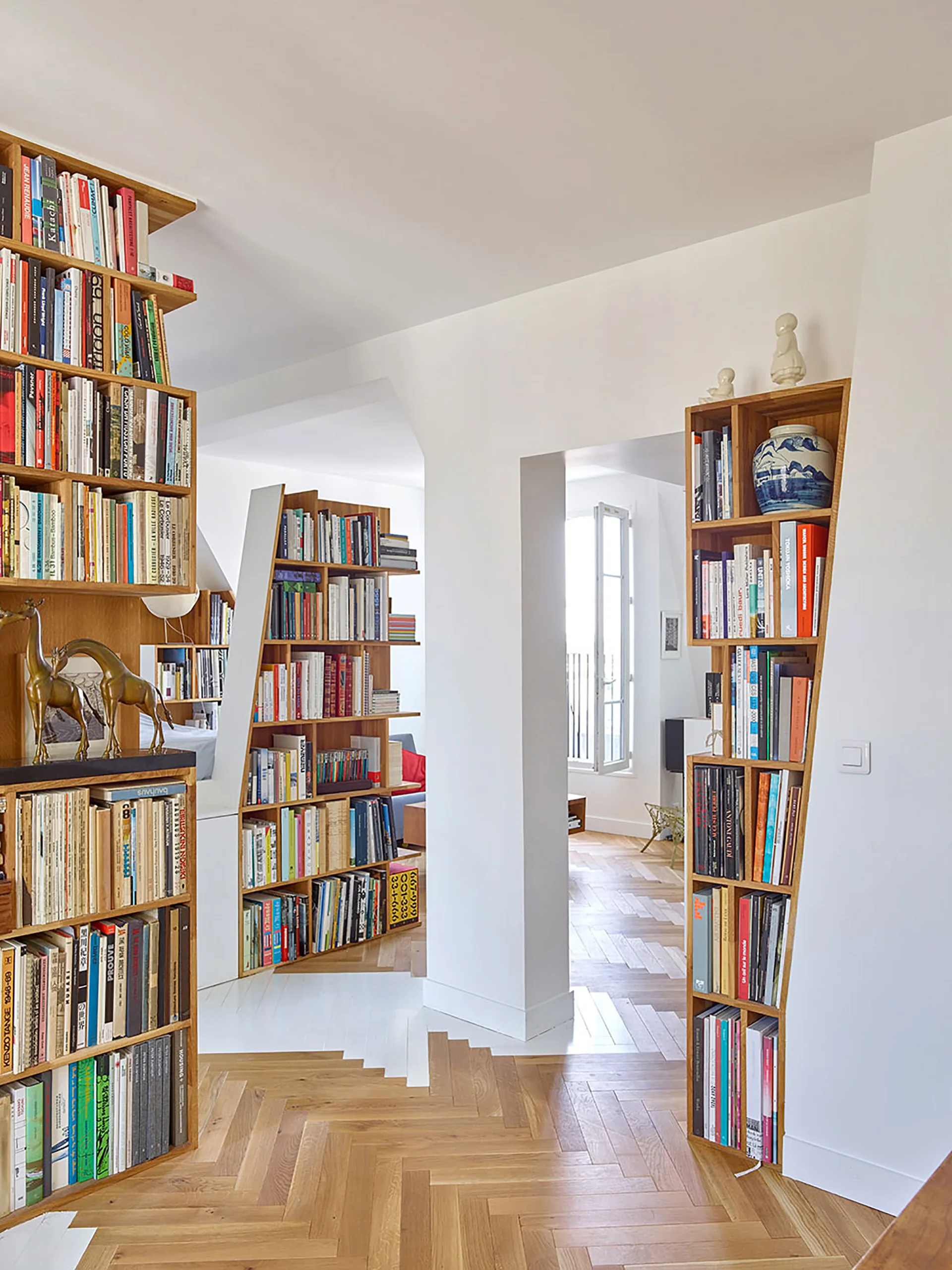 Tall wooden bookshelves filled with books flank a white archway leading to another room with herringbone parquet wood floors.