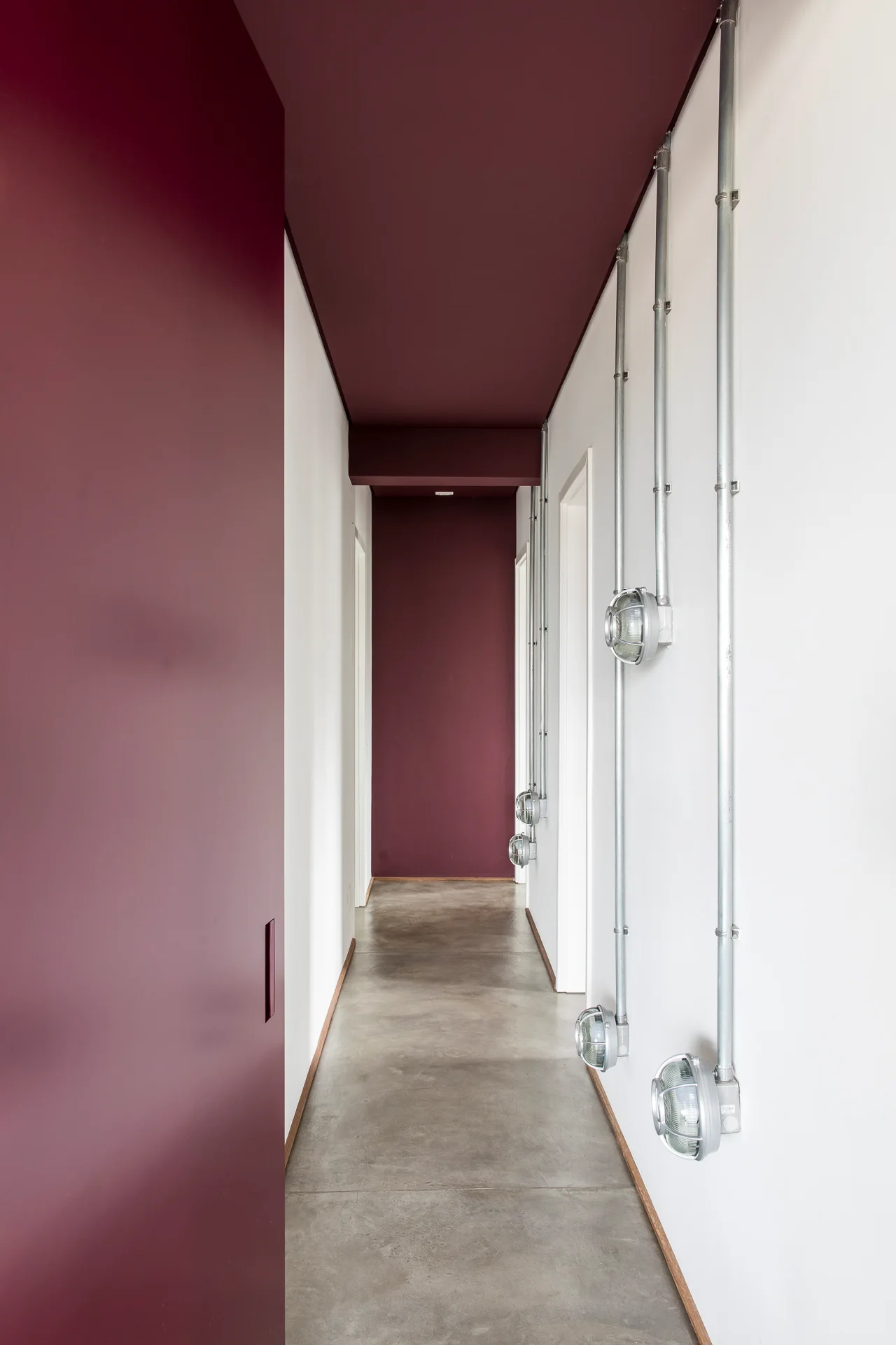 A long hallway features white walls with vertical metal conduits and bulkhead sconces, a grey concrete floor, and a painted deep maroon ceiling.