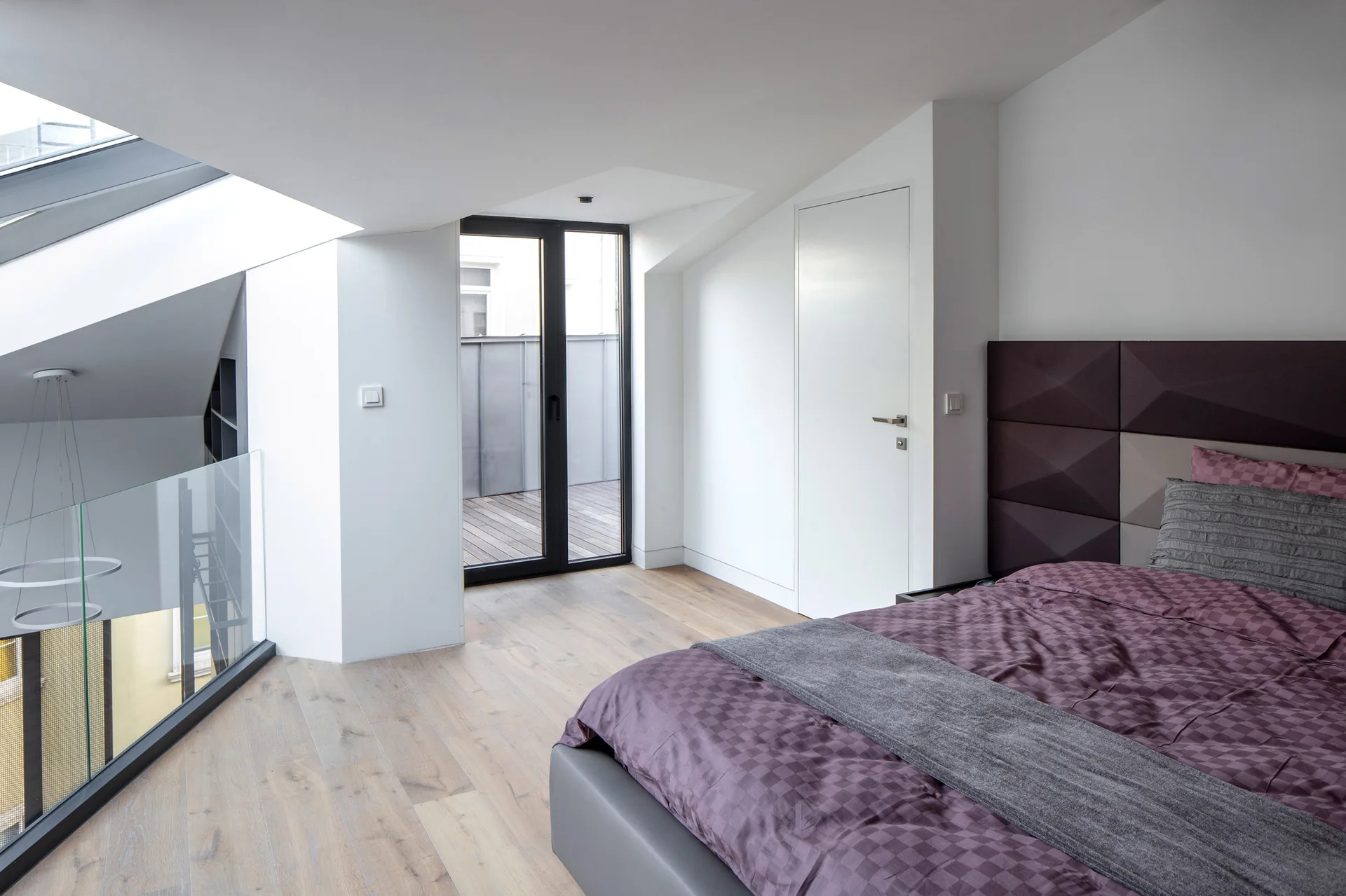 A modern attic bedroom with a bed featuring purple bedding, a large slanted skylight, a glass railing, and a balcony door.