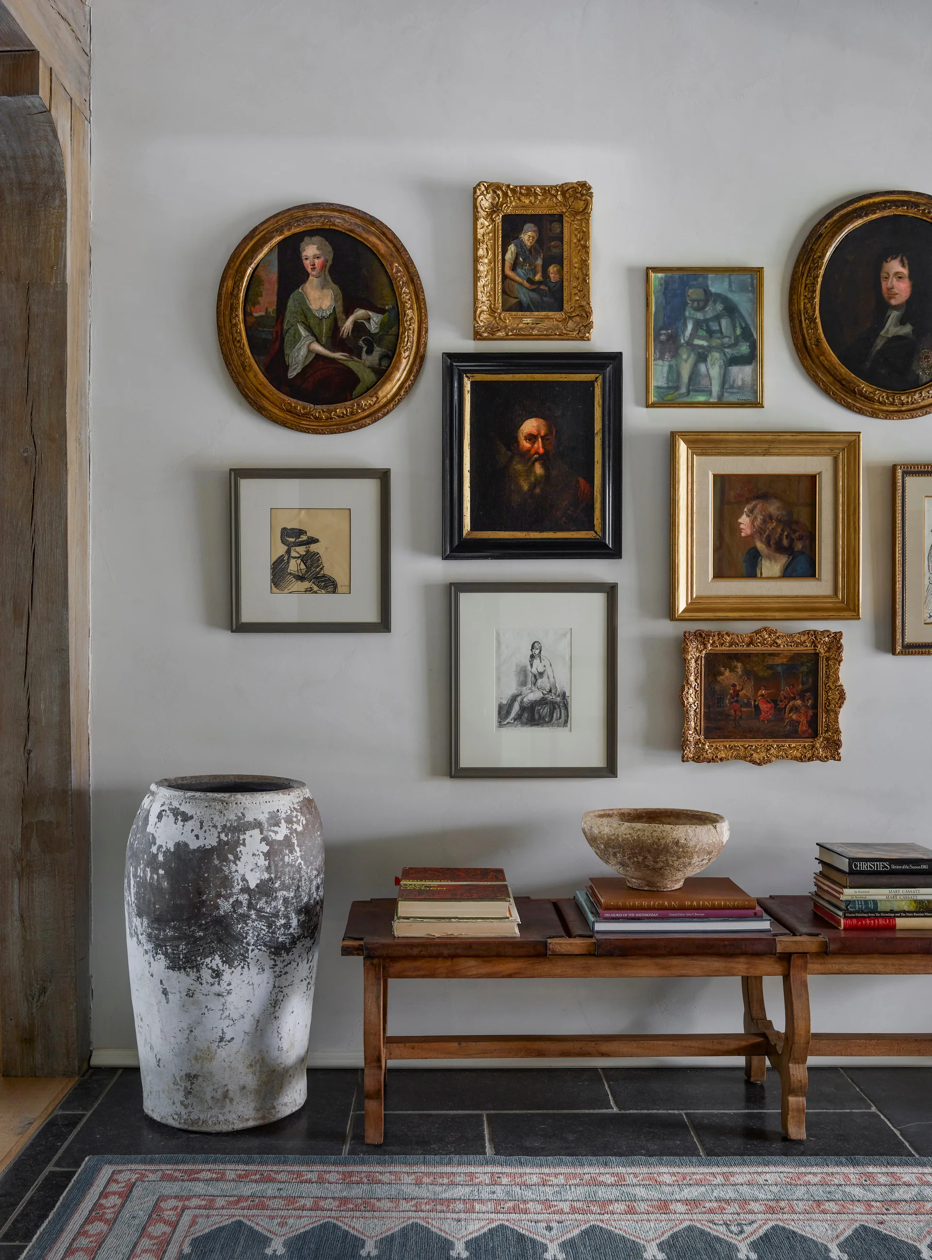 A rustic gallery wall with antique framed portraits hangs above a low wooden bench, a large distressed urn, and dark stone tiles.